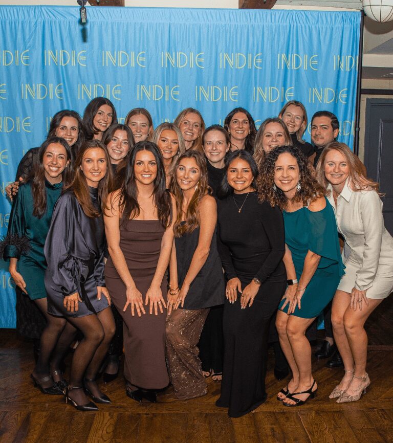 Group of young women posing together in front of a blue backdrop with yellow text, smiling and dressed in formal attire.