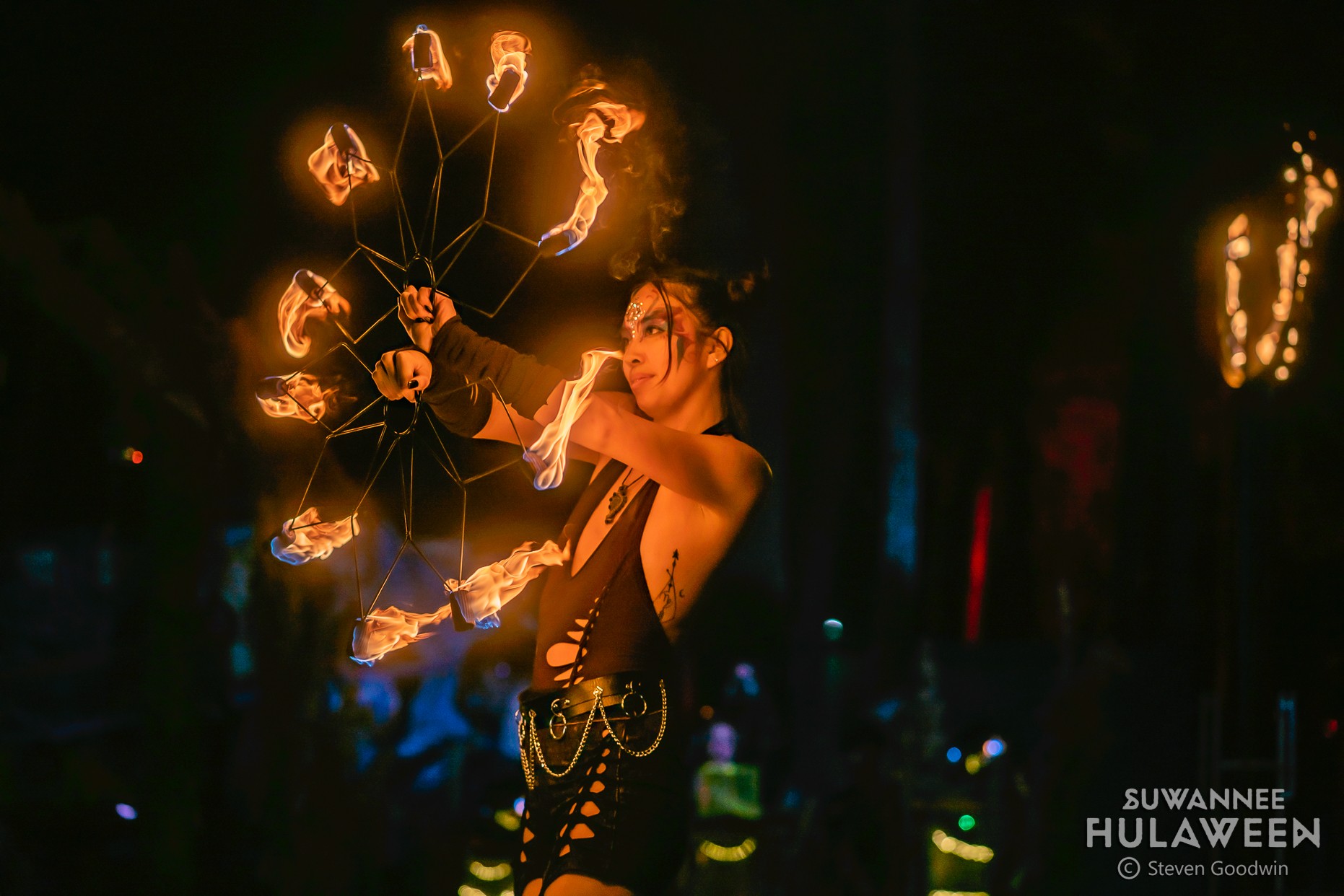 A performer spins flaming poi in a dark setting, with a smiling woman holding fire, surrounded by colorful lights.