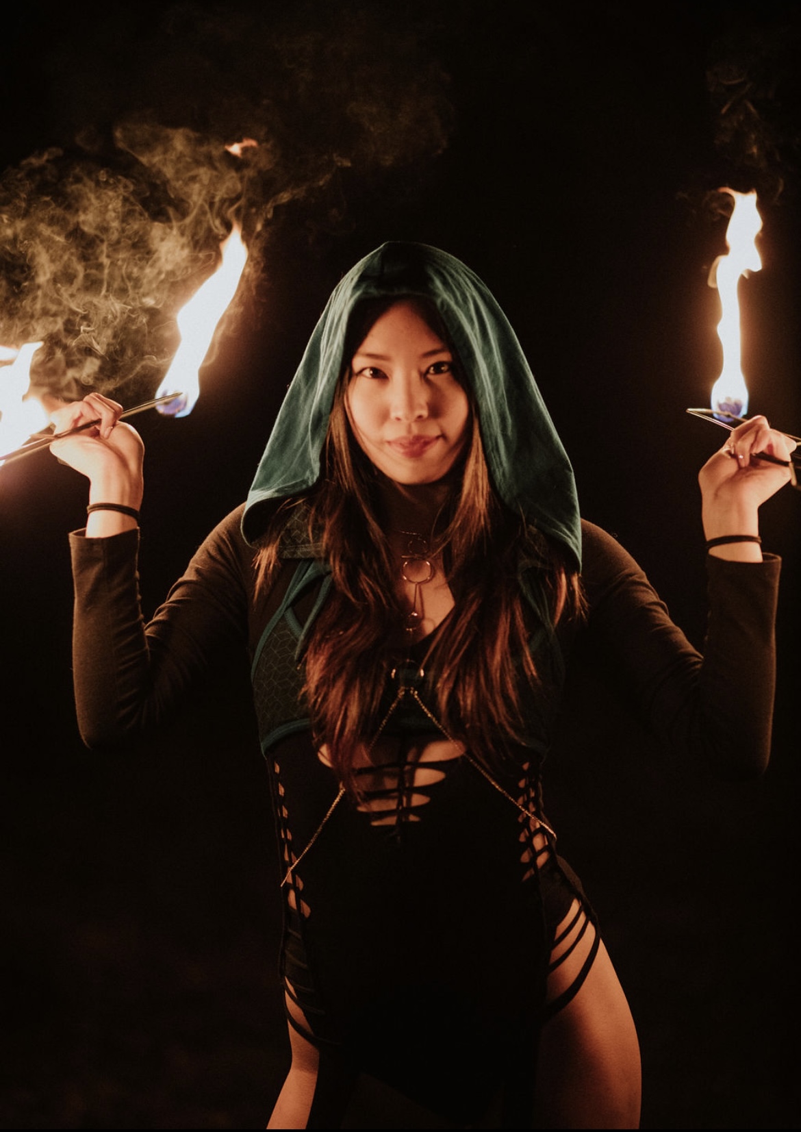 Woman holding fire torches in each hand, wearing a hood and black outfit, dark background, flames visible.