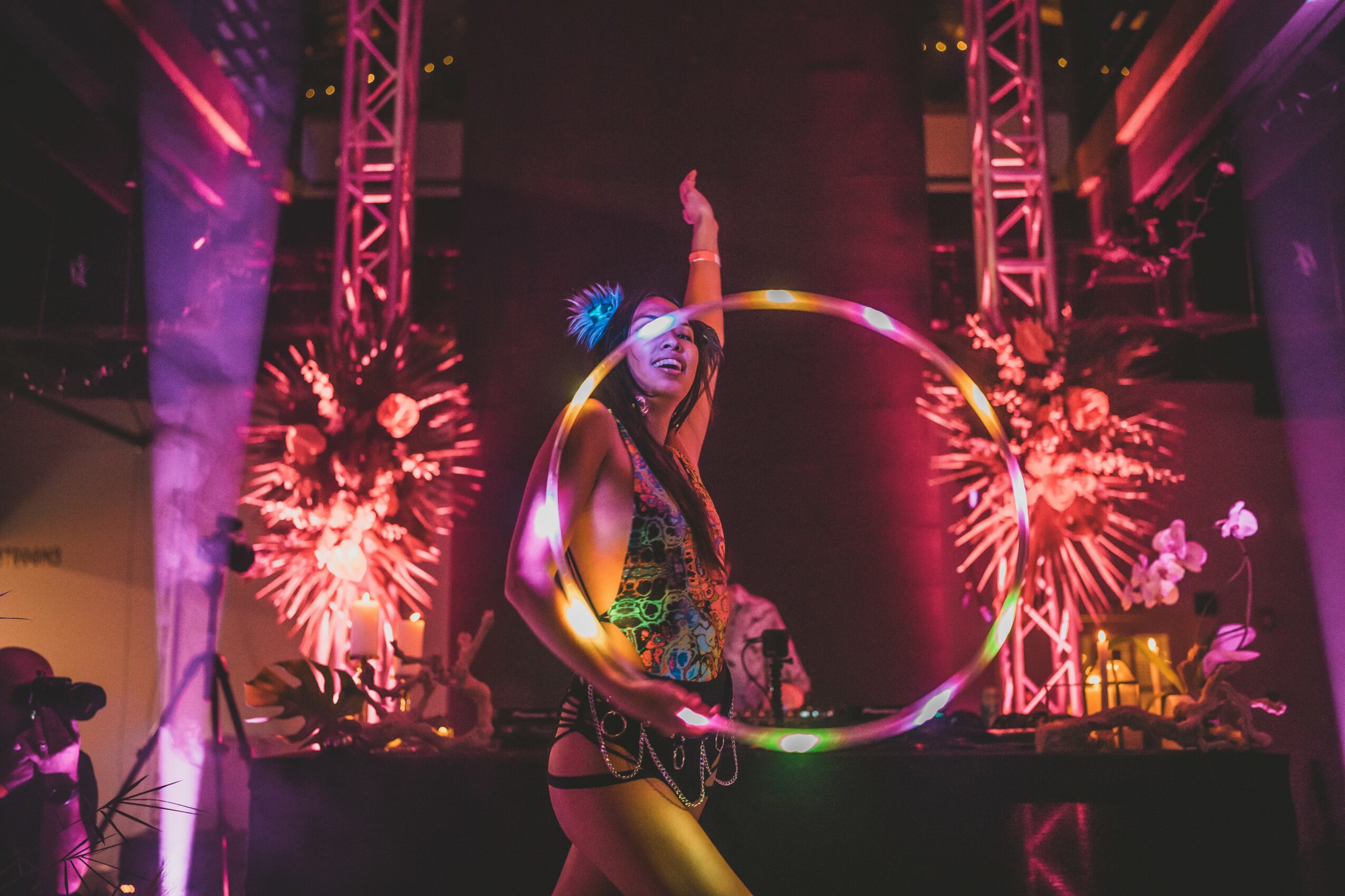 A woman performs with a hula hoop on stage, surrounded by fireworks and colorful lights, in a lively event setting.