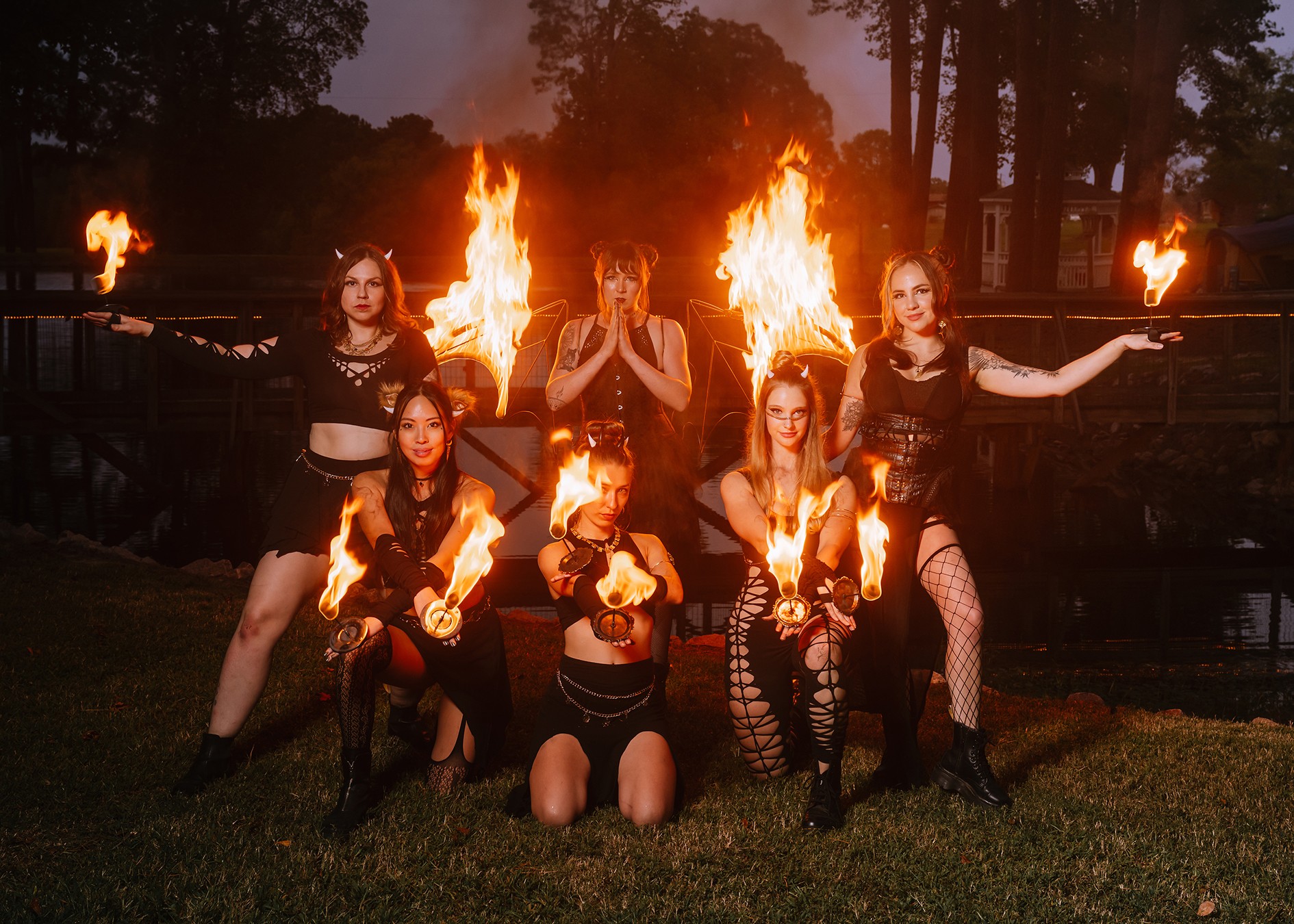Six women perform a fire dance outdoors at night, with flames behind and around them, some holding fire torches.