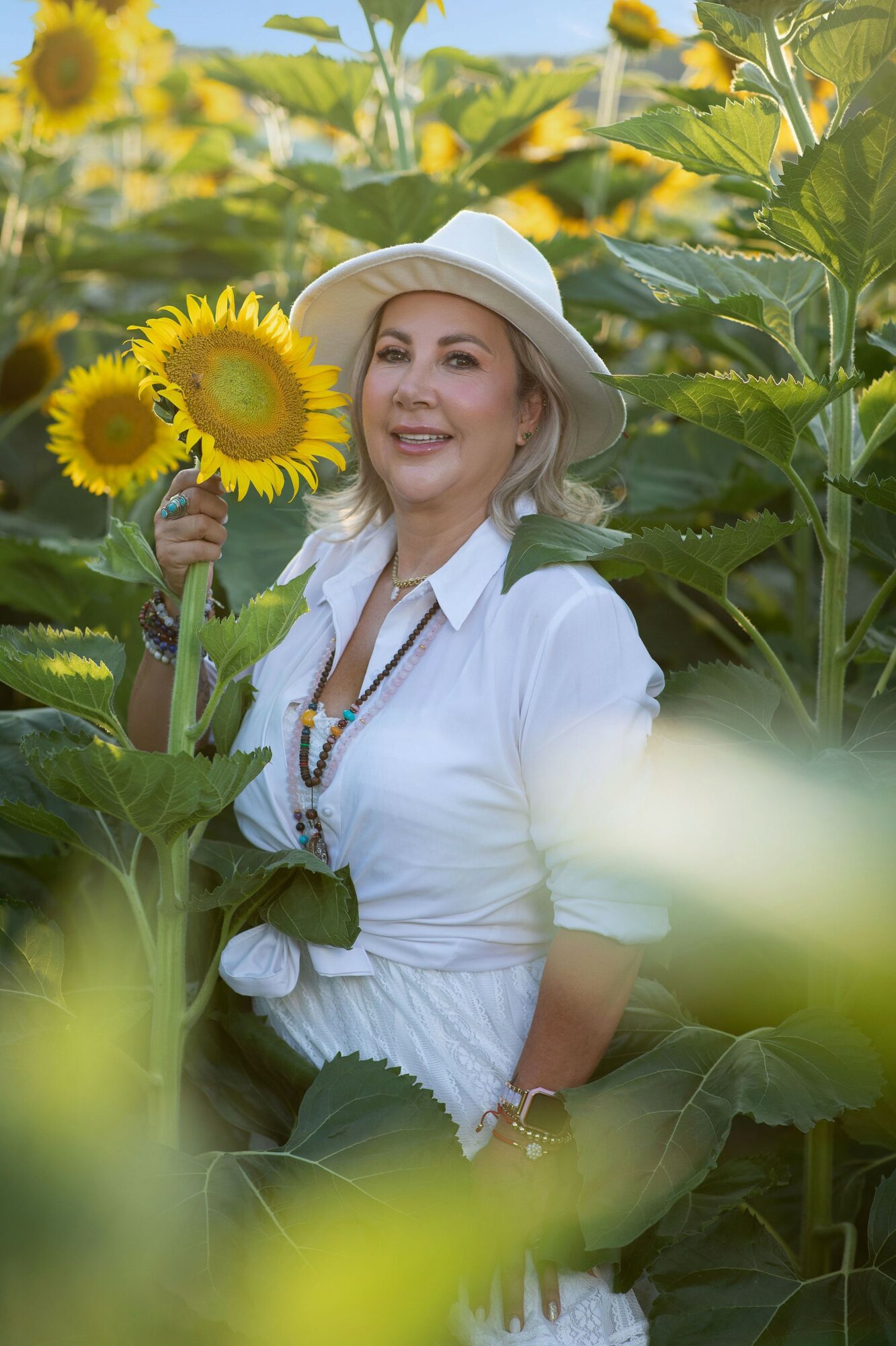 Woman in white shirt and hat holding sunflowers in a sunflower field, smiling, sunlight, green leaves, bright scene