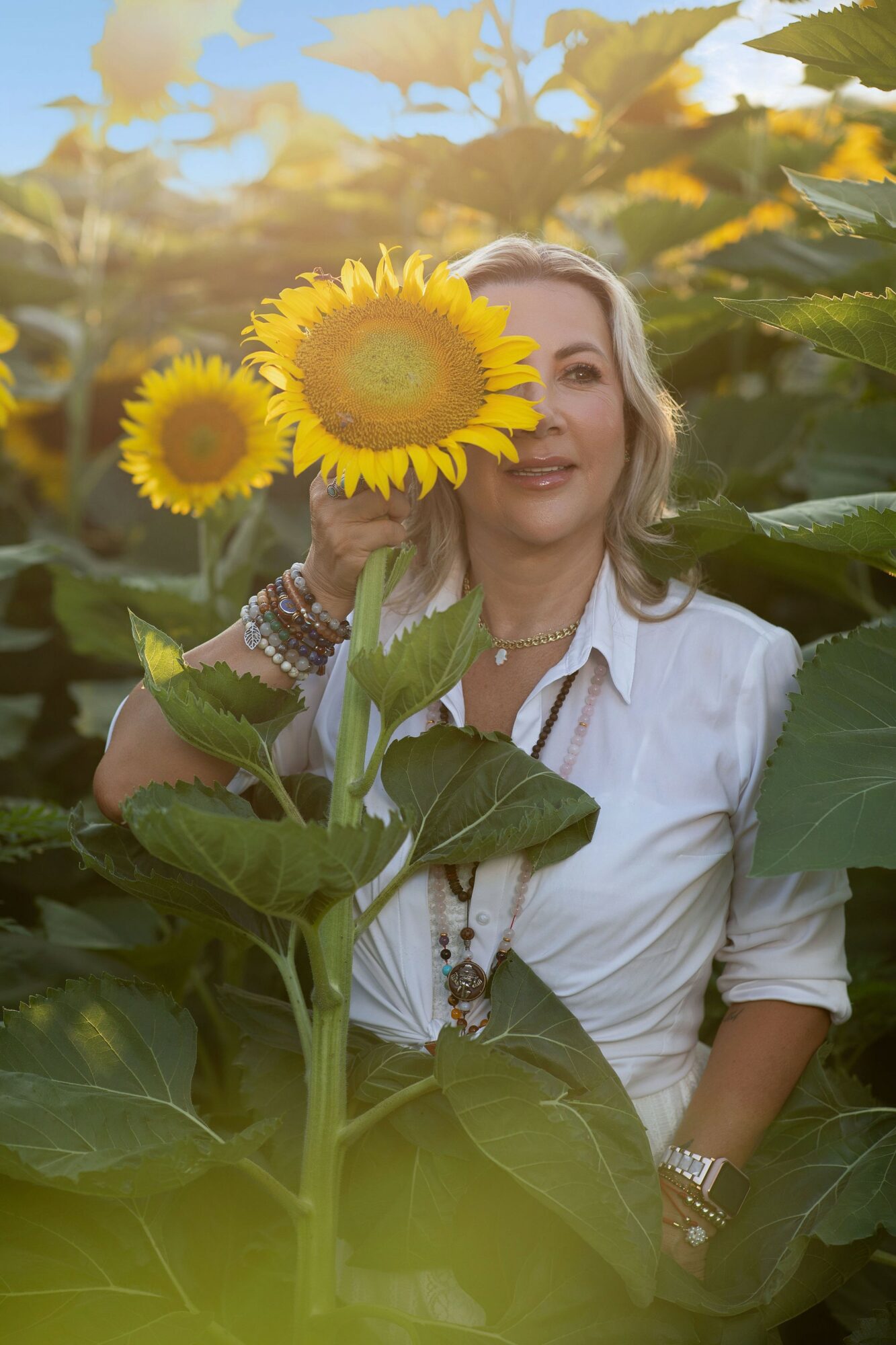 Woman with blonde hair holding a sunflower in a sunflower field, smiling, wearing a white shirt and jewelry.