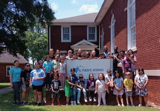Group of children and adults standing outside a brick building holding a sign that reads 'El Puente Hispano'.