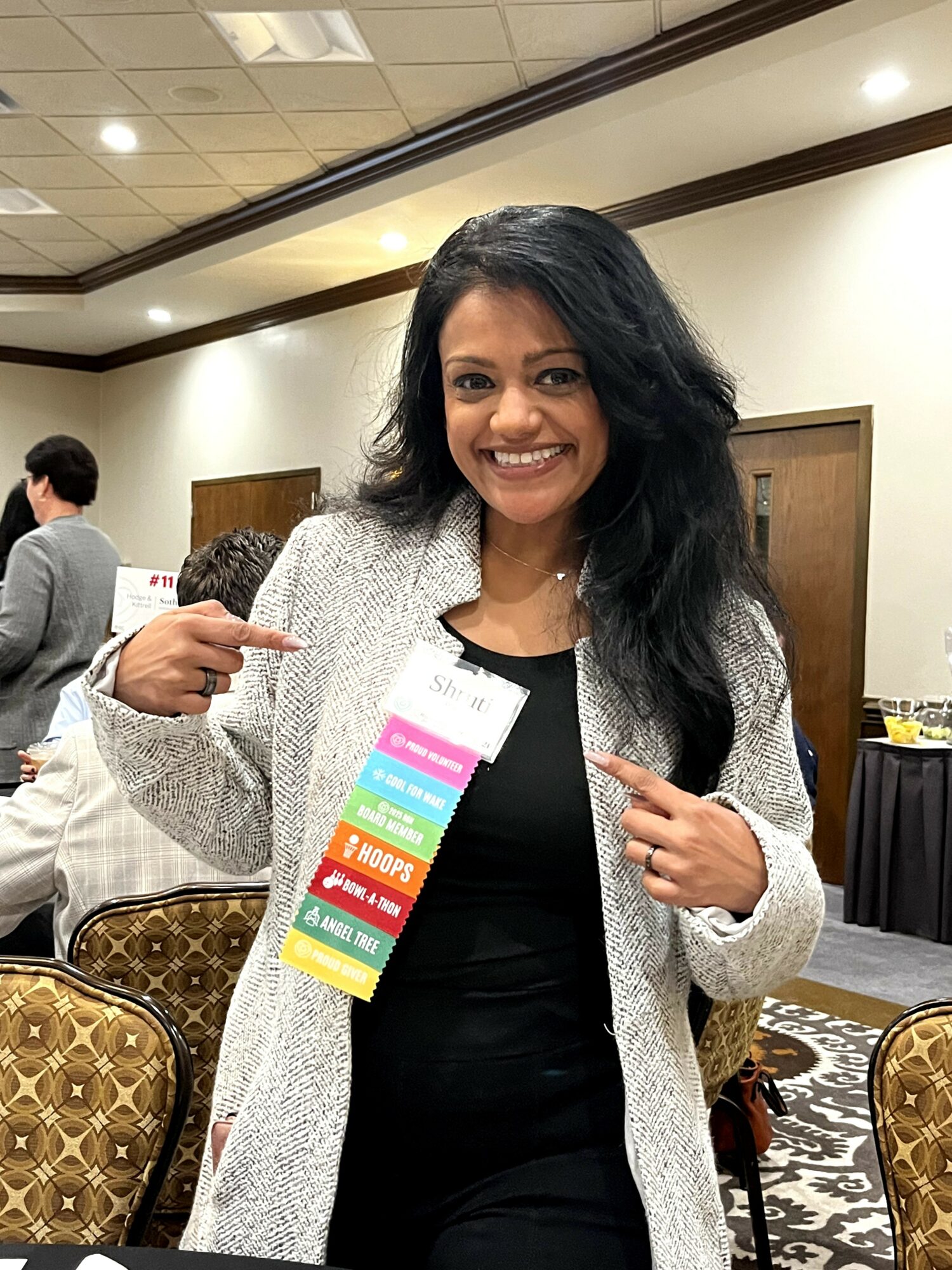 Smiling woman pointing at a colorful badge, standing in a conference room with chairs and other people.