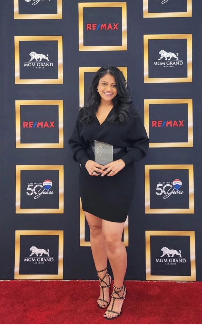 Woman in black dress holding an award, standing on red carpet in front of a backdrop with logos and text.