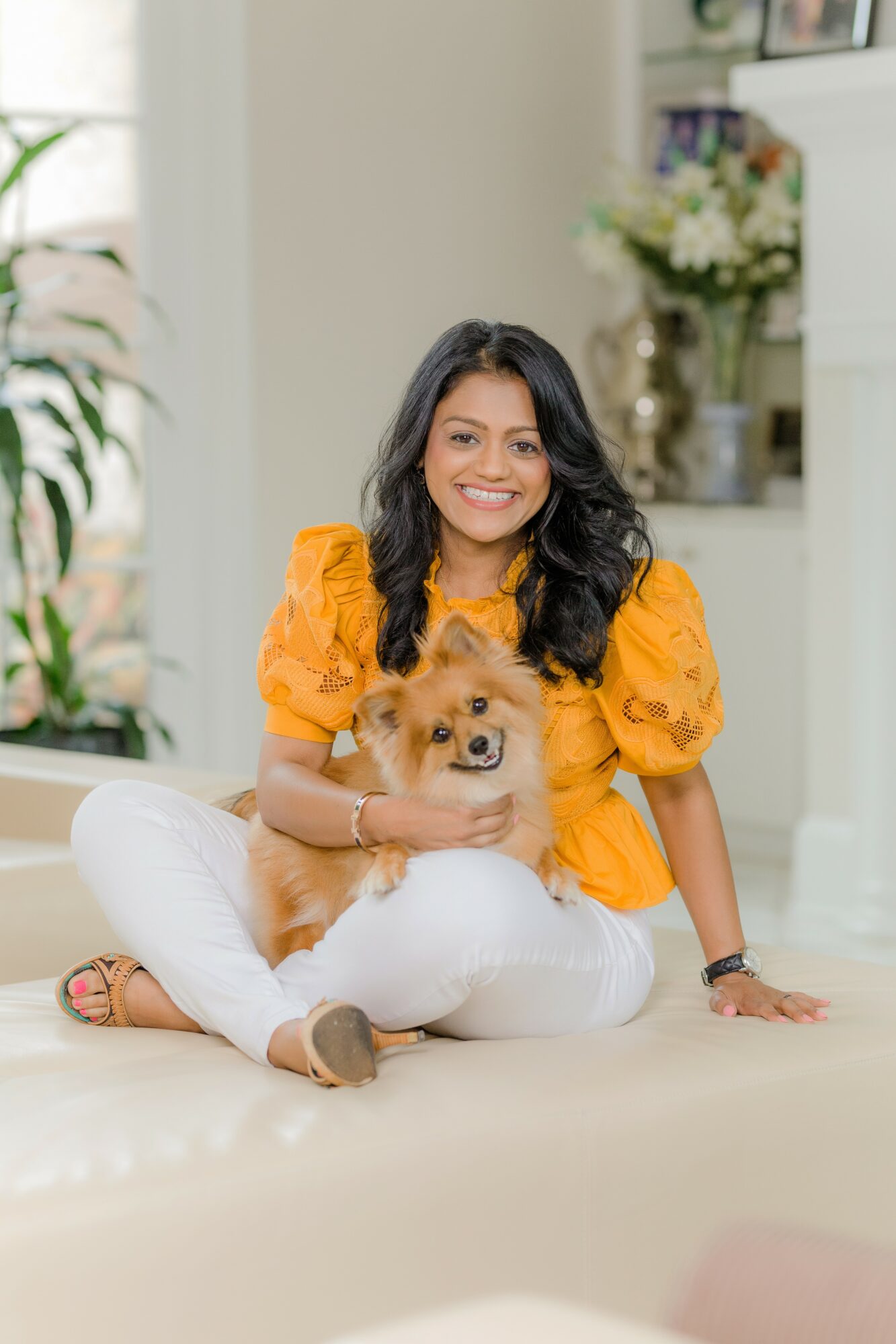 Woman in yellow top sitting on floor with small dog, smiling, in bright room with plants and flowers