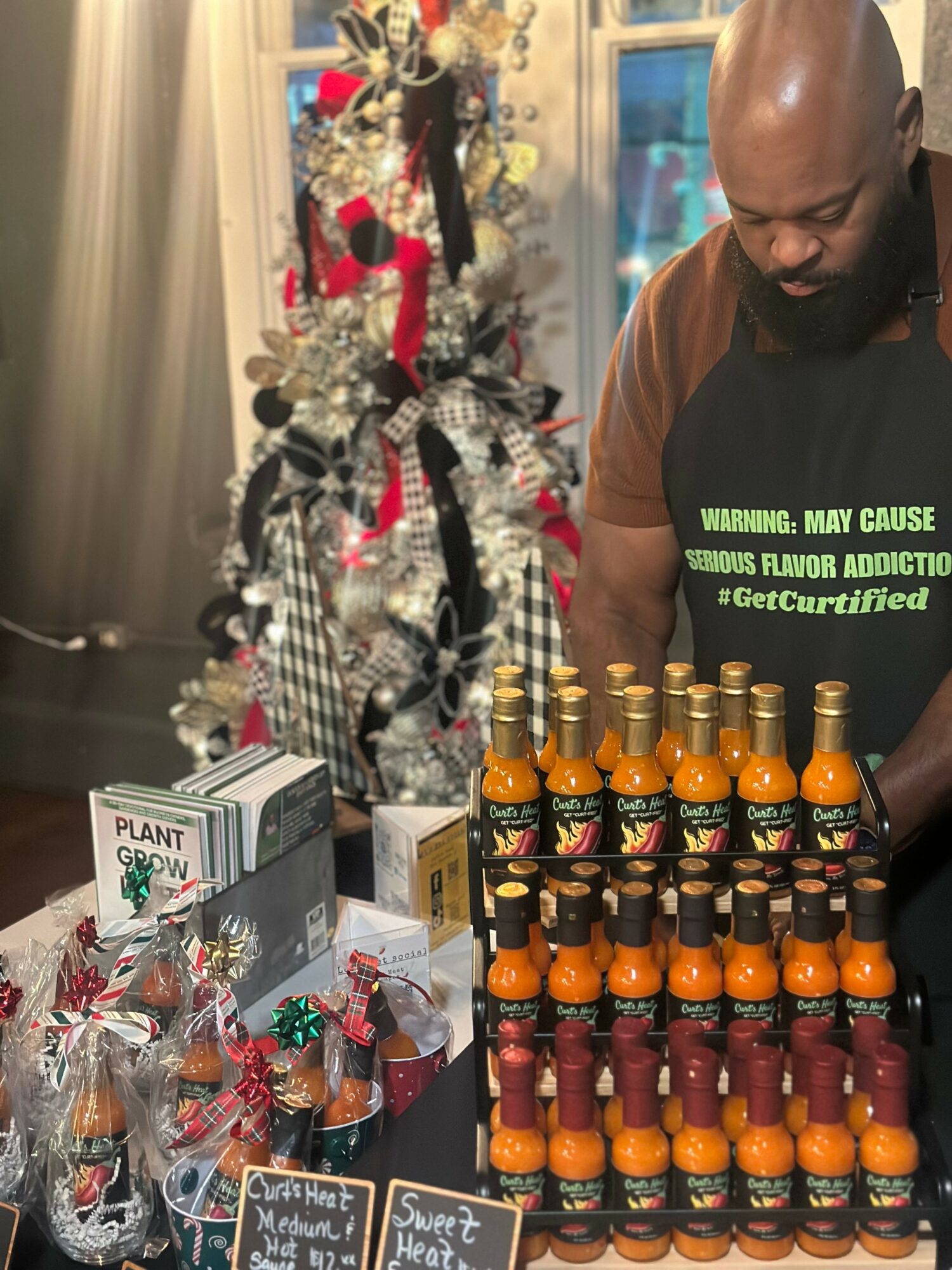 Man with a beard looking down at a display of small bottles, Christmas tree decorated with ribbons and ornaments in background.