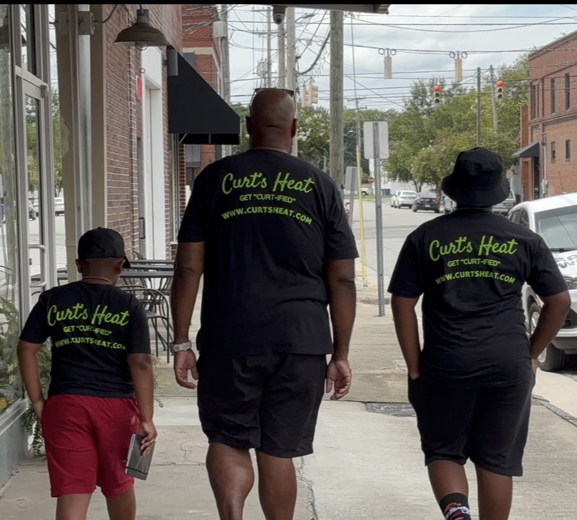 Three people walking on a sidewalk, wearing black shirts with green text, in an urban area with buildings and power lines.
