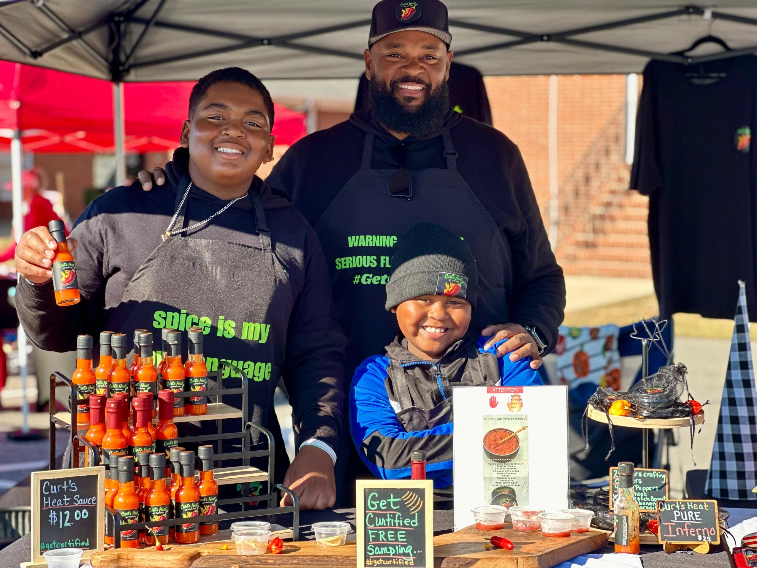 Two smiling people and a child behind a booth with hot sauce bottles, signs, and merchandise outdoors under a canopy.