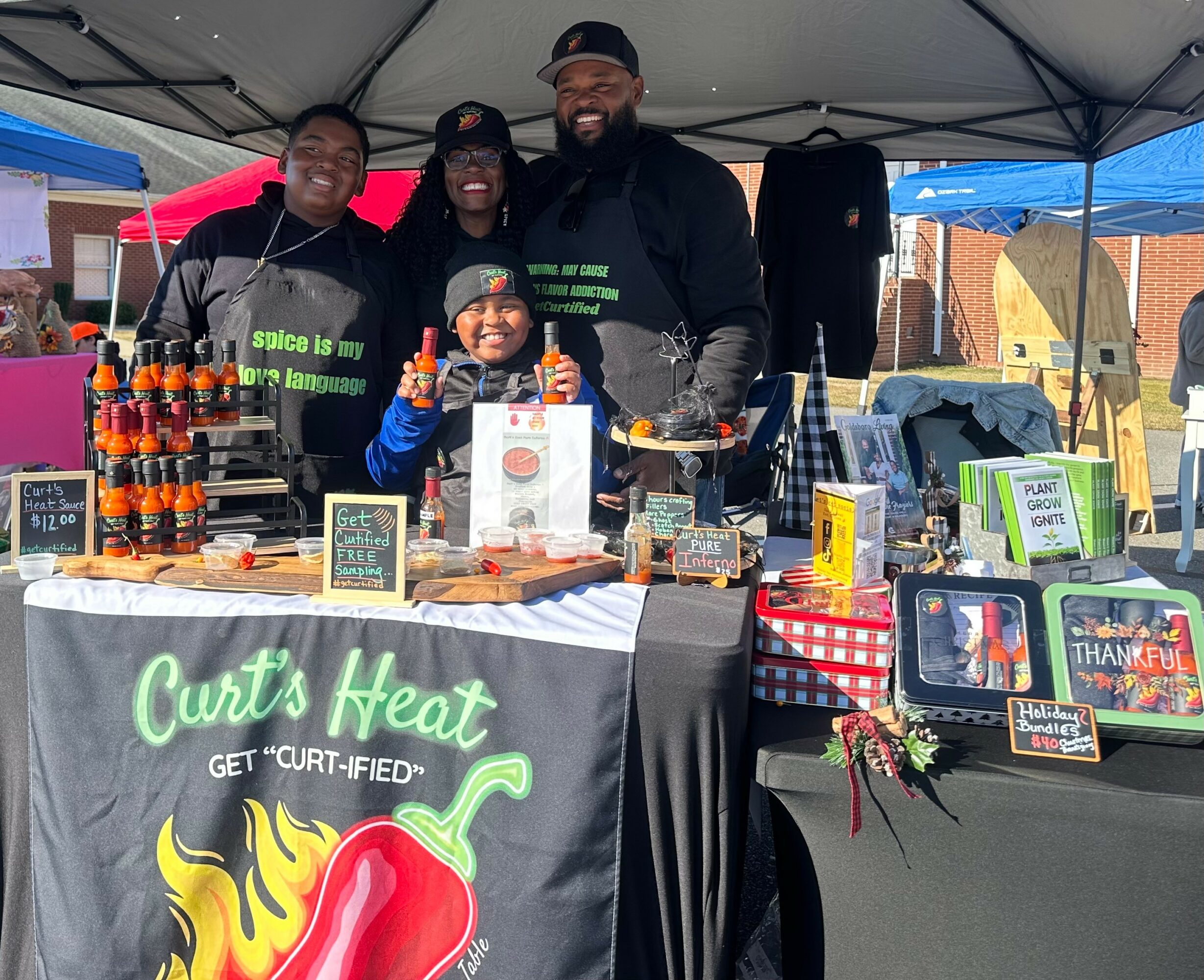 Four people stand behind a table at an outdoor market stall, smiling, with various products displayed on the table.