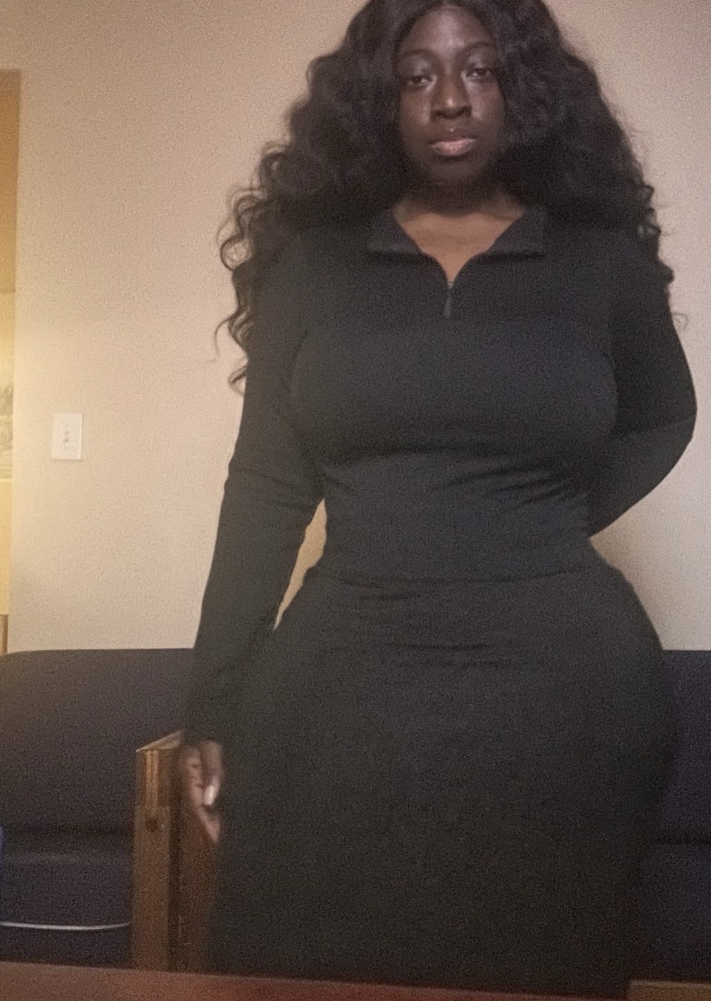 Woman with long curly hair wearing a black dress standing indoors against a beige wall.
