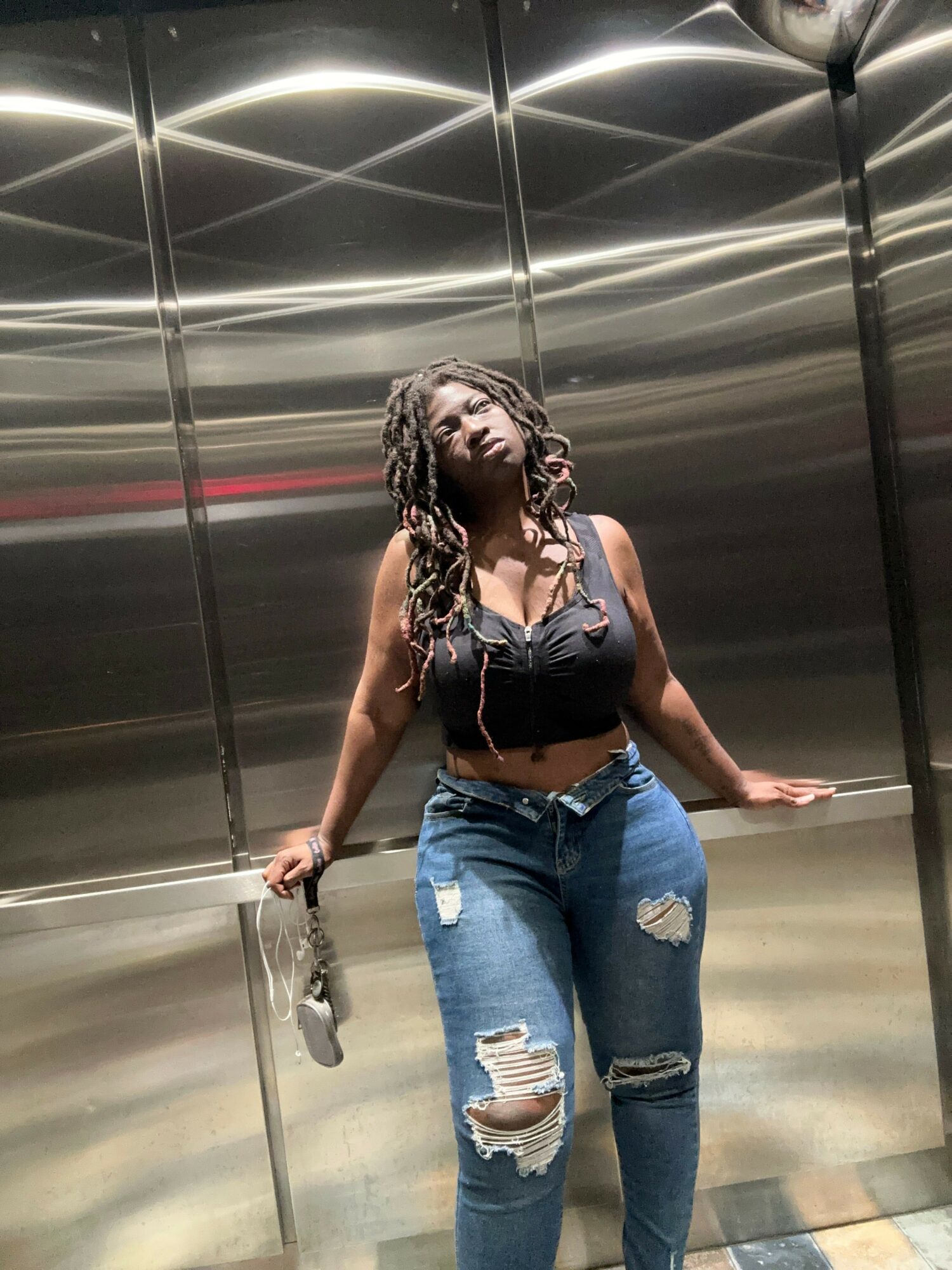 Woman with long curly hair in ripped jeans and black top standing in an elevator, holding a phone.