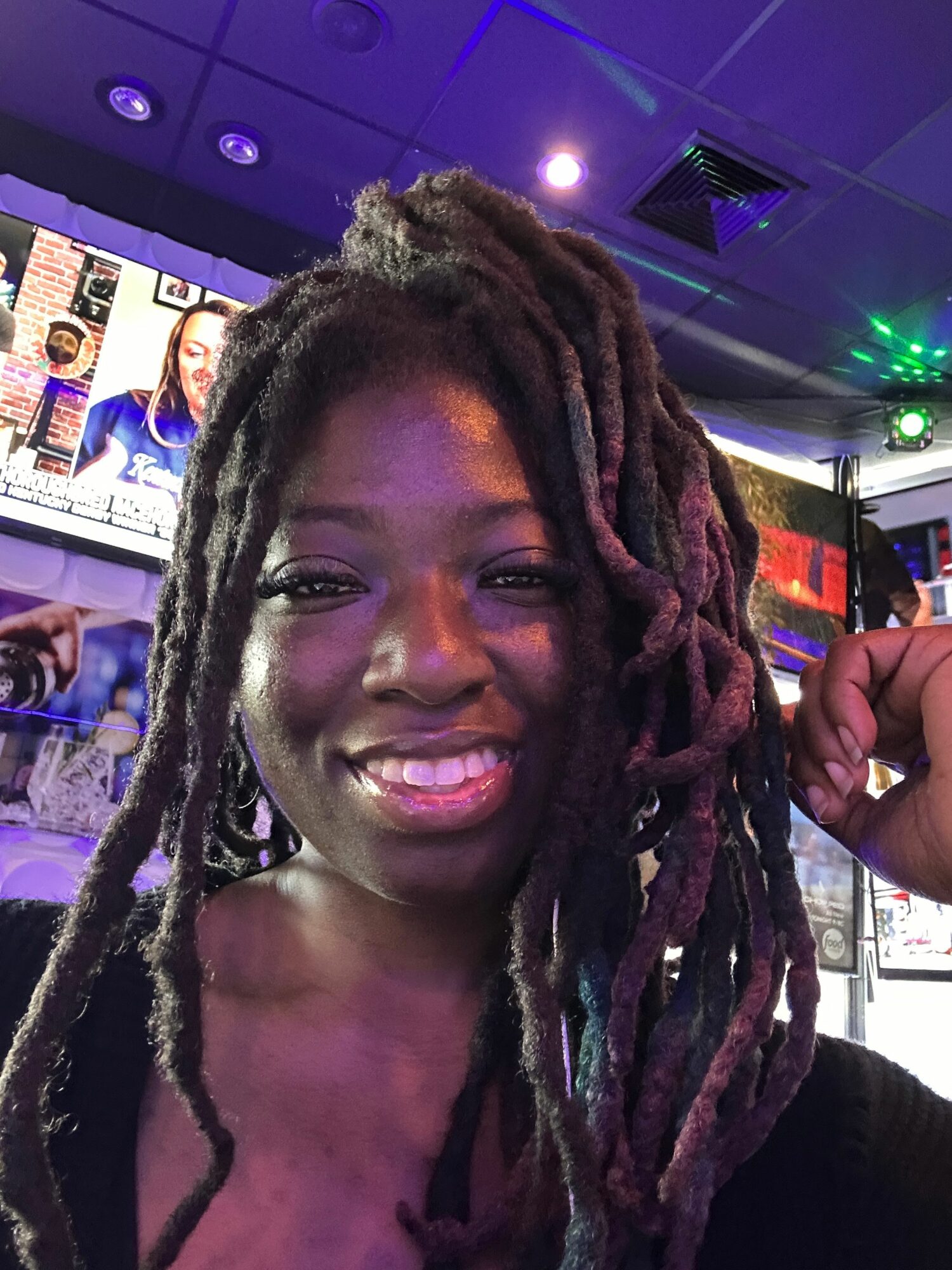 Woman with dreadlocks smiling in a lively indoor setting, colorful lights and screens in background.