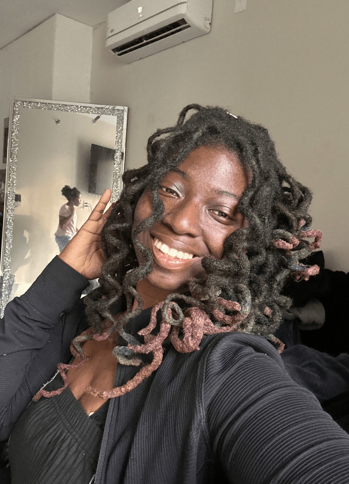 Smiling woman with styled curly hair taking a selfie in a room with a mirror and air conditioner visible.