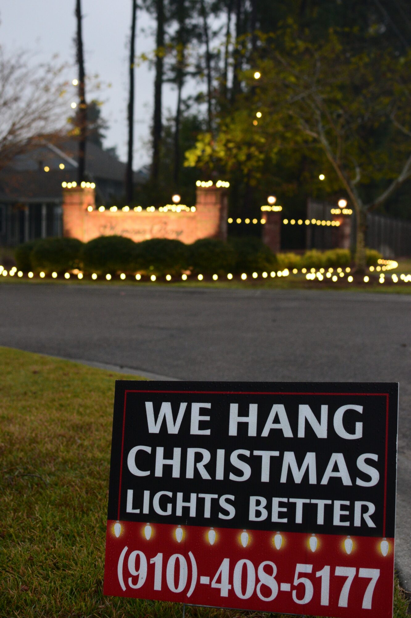 Christmas lights decorate a fence and trees in a yard at dusk, with a sign in the foreground reading 
