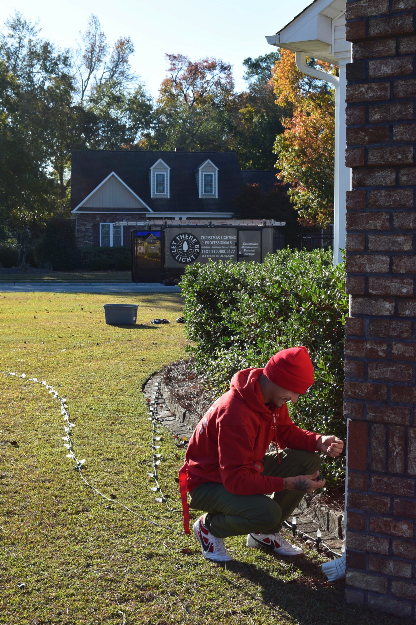 Child in red coat and hat crouching near brick wall, working on a project outside, with house and trees in background.