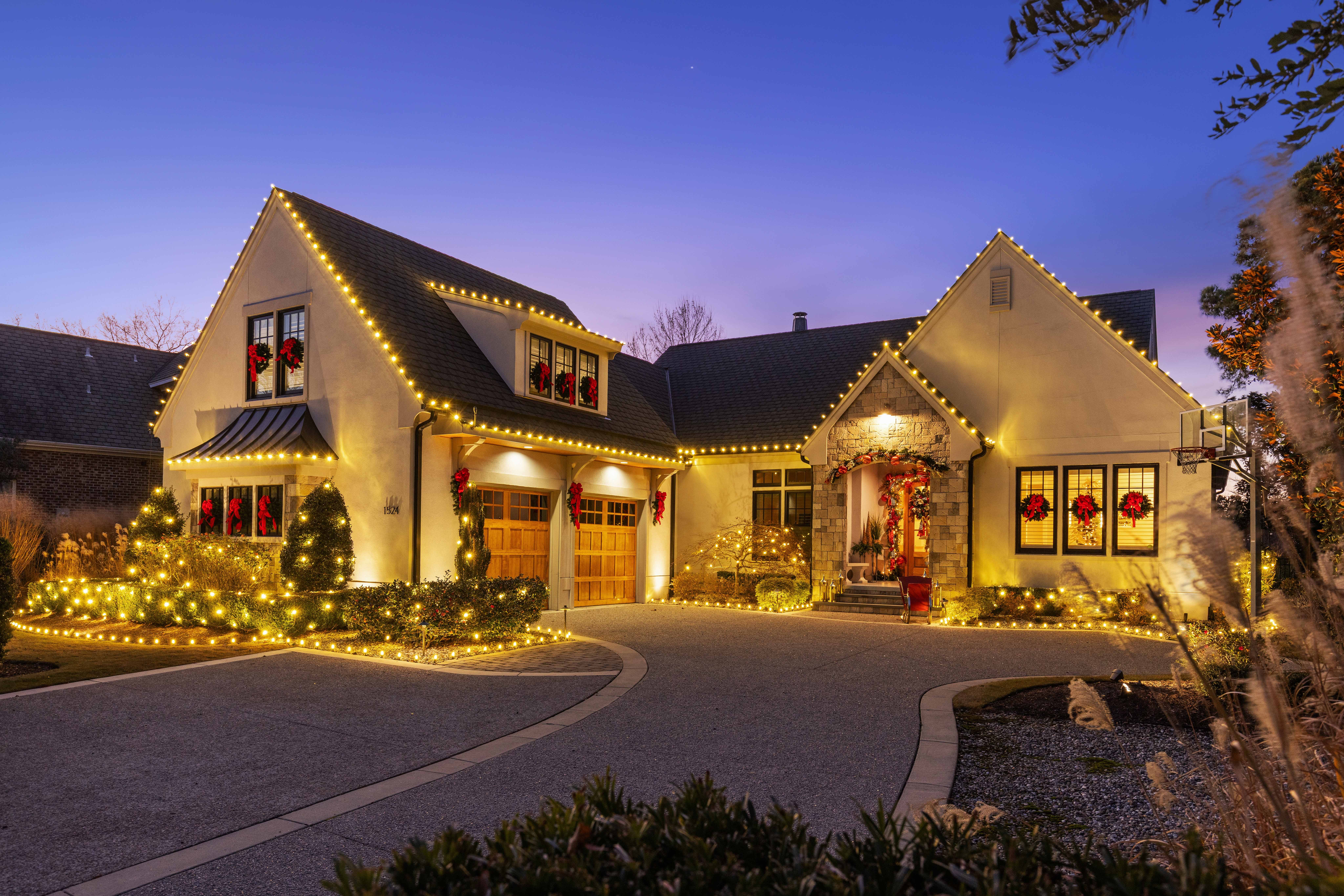 House decorated with Christmas lights and wreaths, illuminated at dusk, with a curved driveway and landscaped yard.