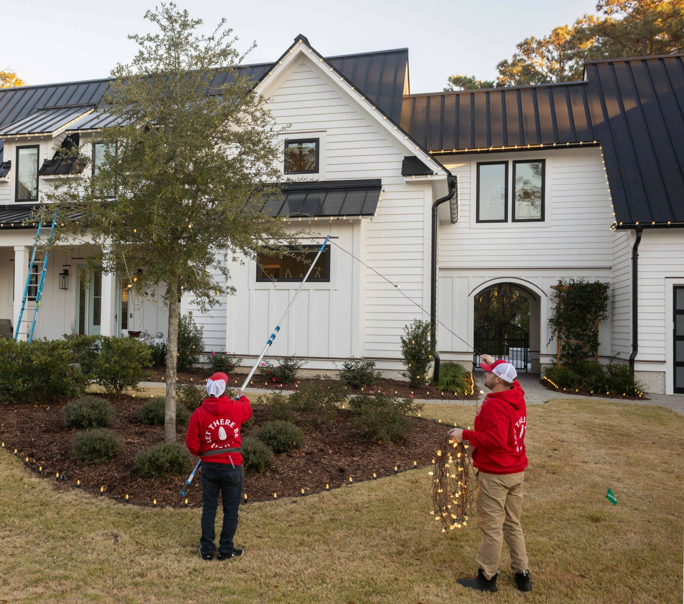 Two children in red jackets and Santa hats stand in front of a white house with black roof, holding a string of lights.