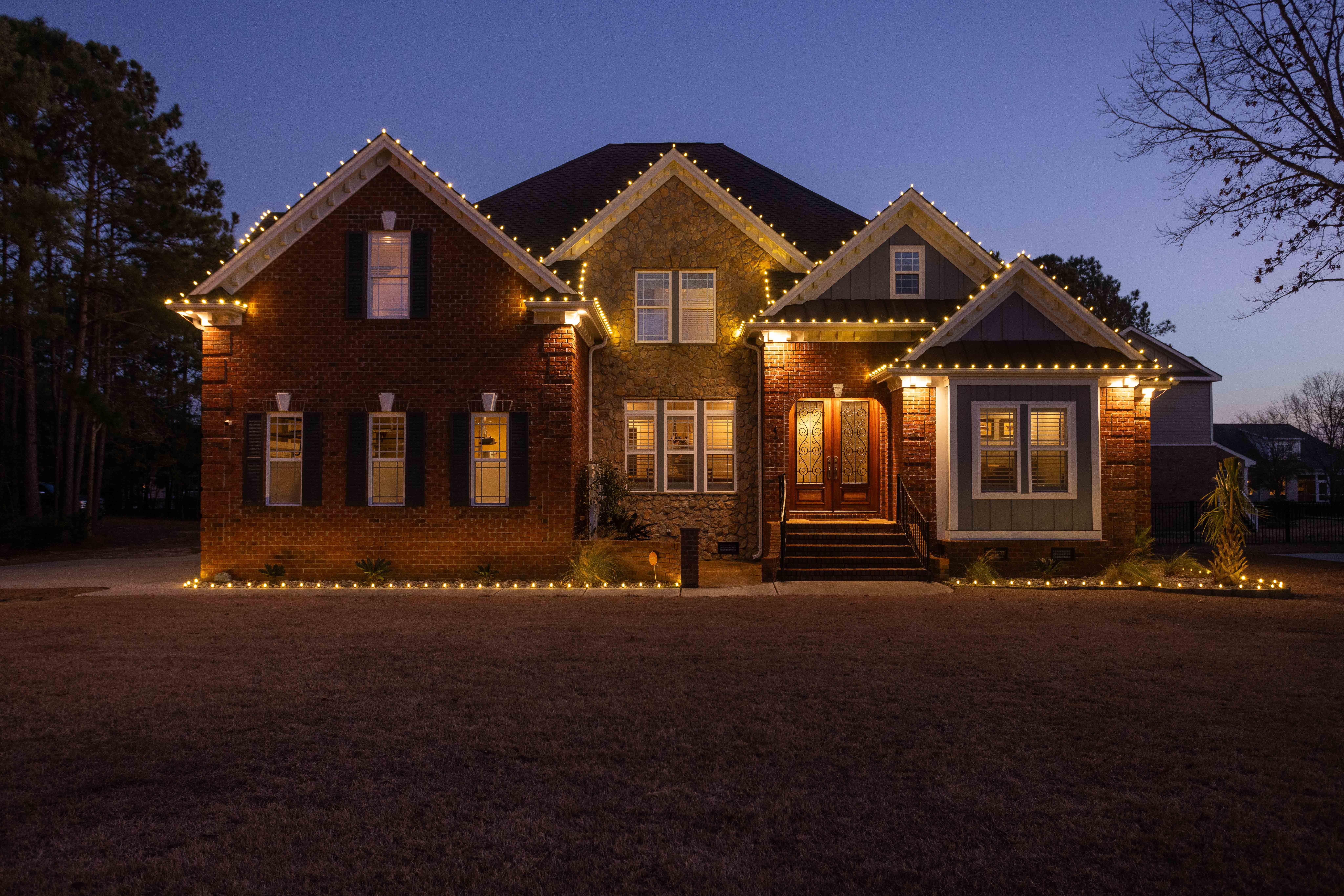 Large house with multiple gabled roofs, illuminated windows, and a front porch, set against a twilight sky.