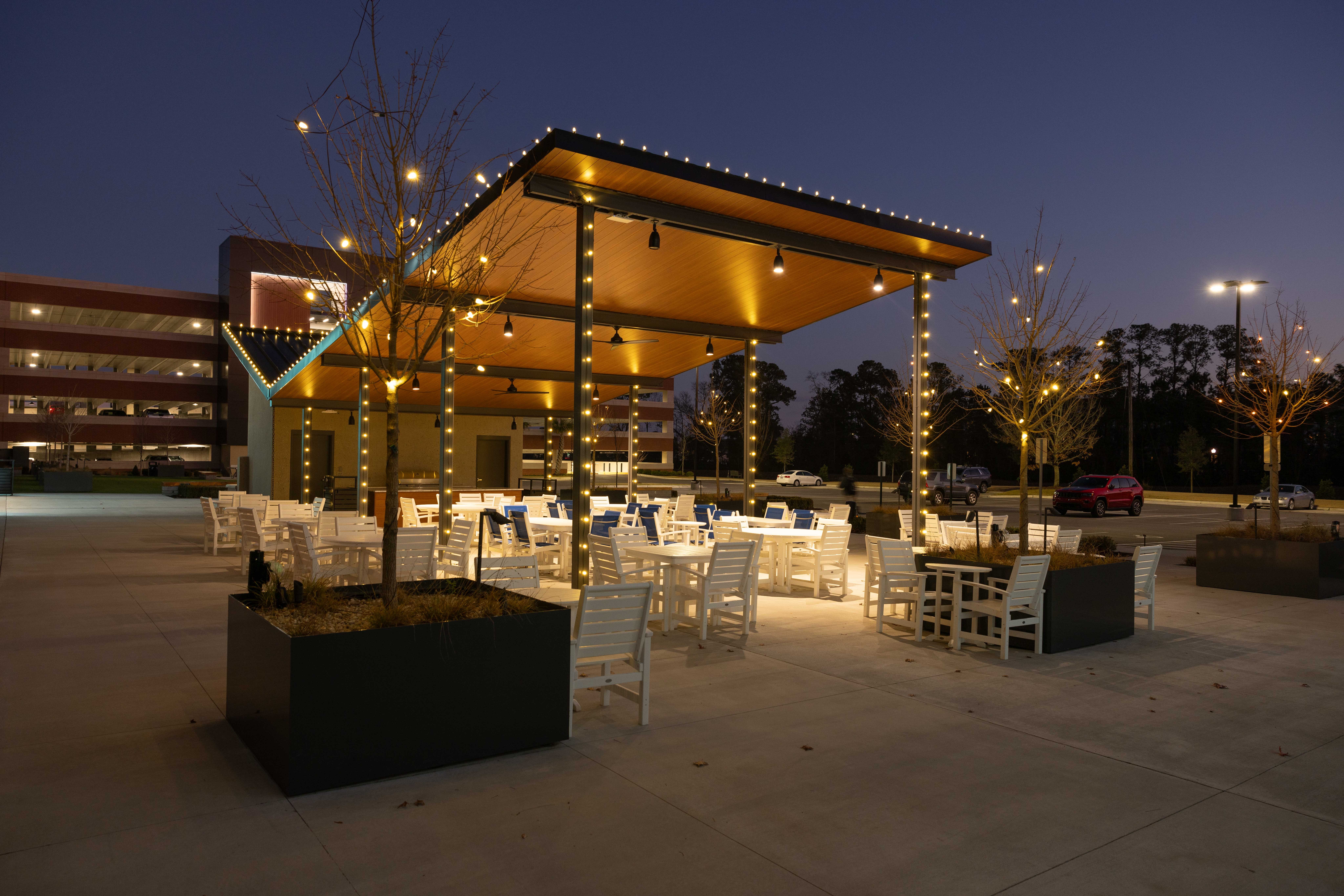 Outdoor seating area with tables and chairs under a lit canopy at dusk, trees and buildings in background.