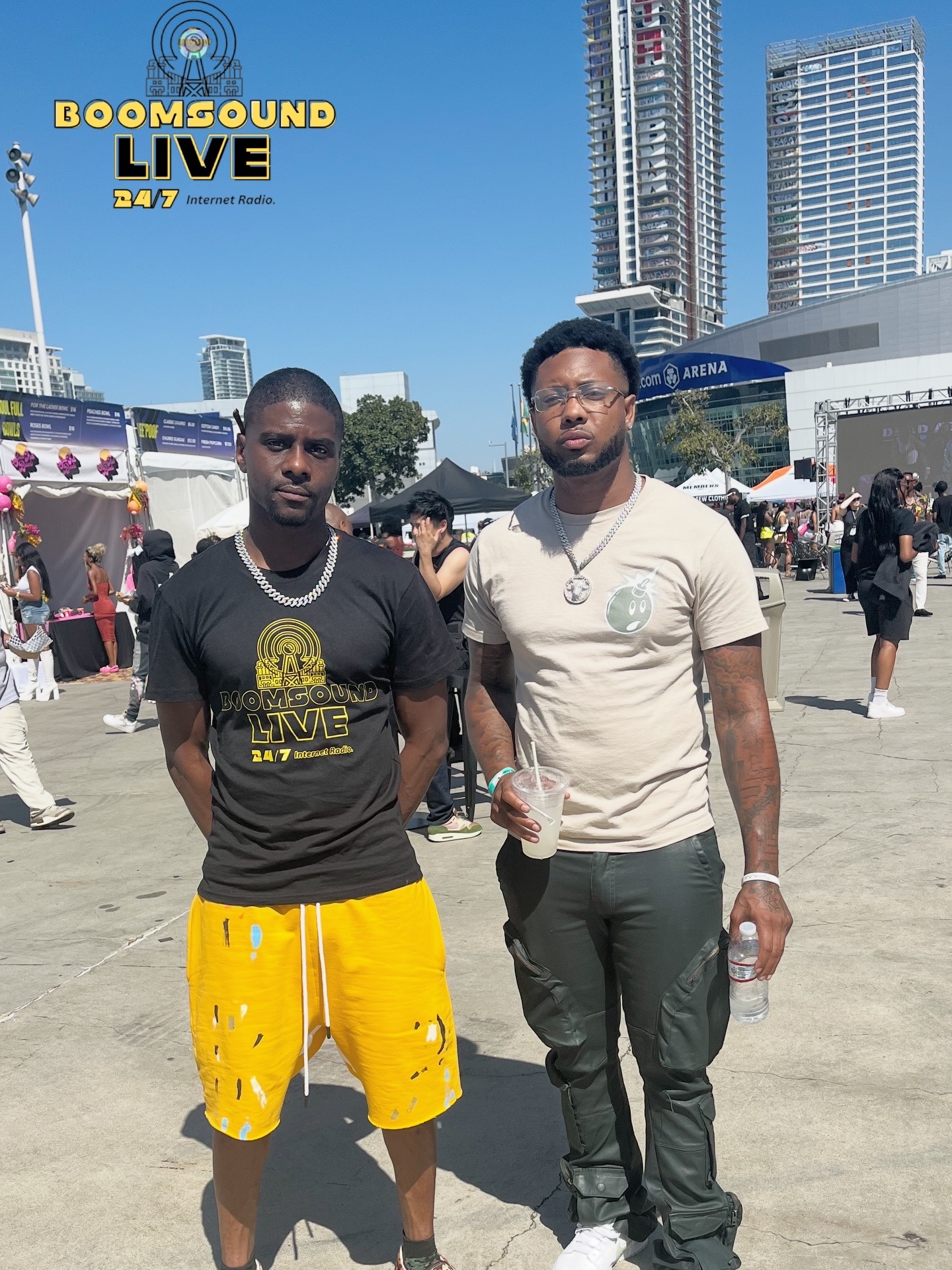 Two men stand outdoors in a city square with tall buildings and a crowd, one wearing a black T-shirt and yellow shorts, the other in a beige T-shirt and dark pants, holding a water bottle.