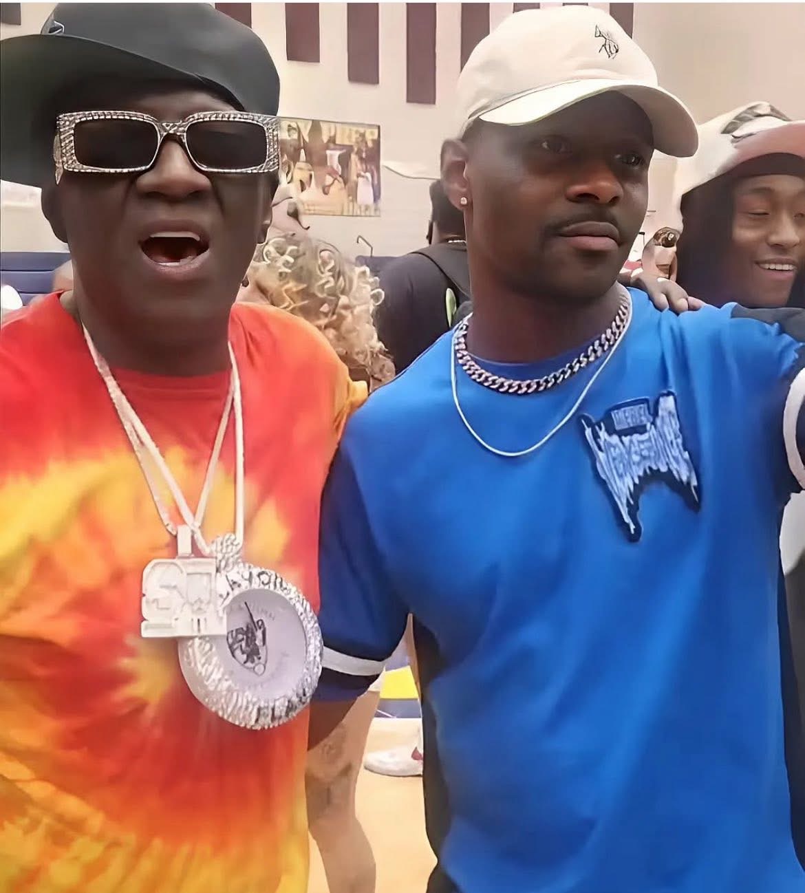 Two men in colorful clothing and accessories, one wearing sunglasses and large jewelry, the other in a blue shirt and white cap, smiling.