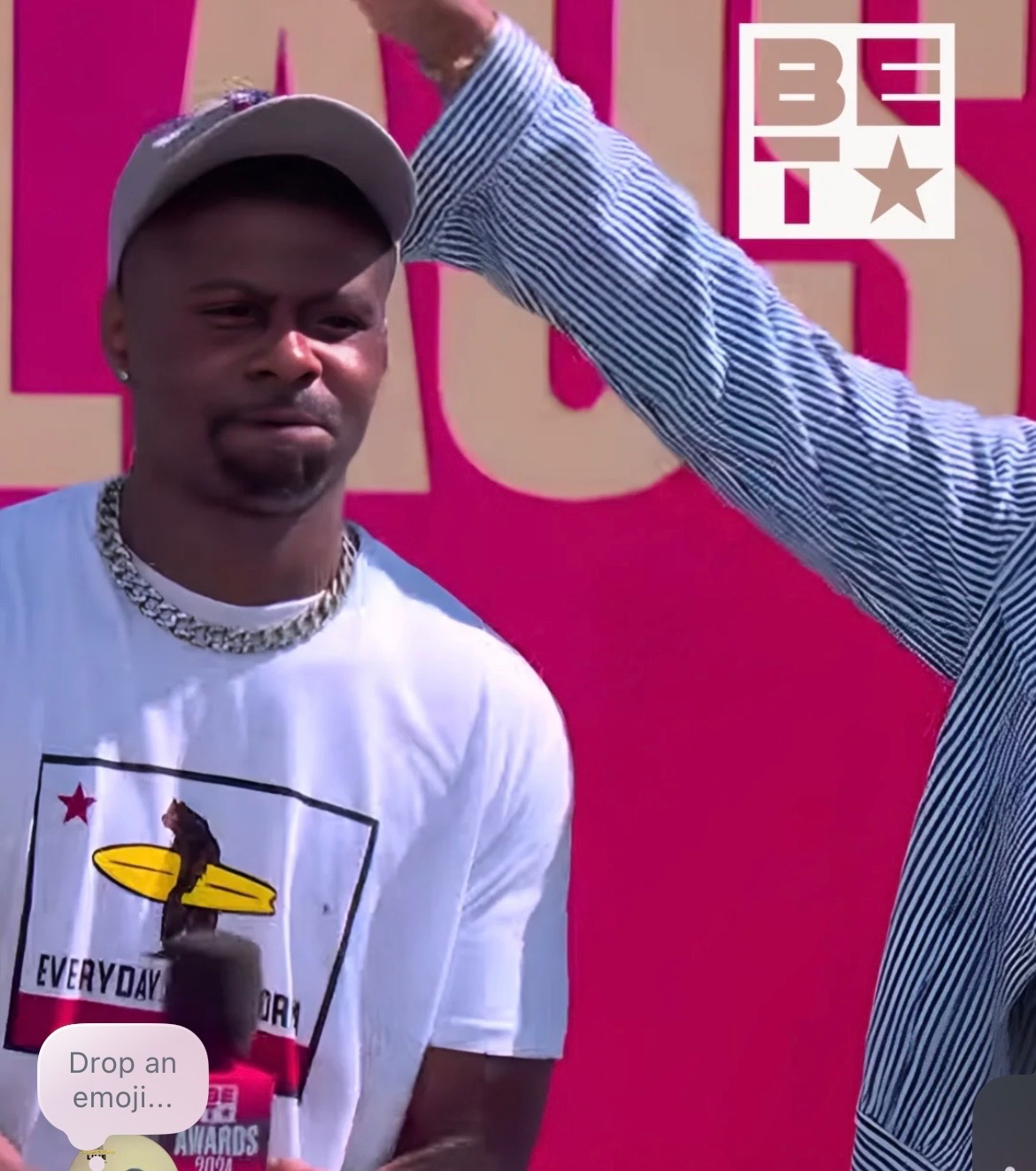 Young man with a cap and chain, standing outdoors with a pink background, another person's arm adjusting his cap.