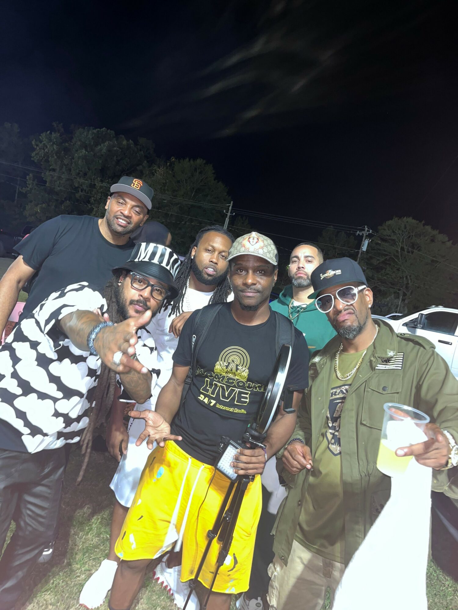 Group of six people outdoors at night, some holding drinks, smiling, with trees and a building in background.