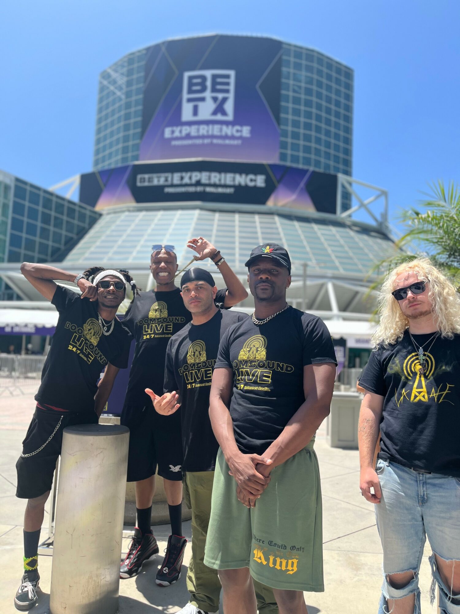 Group of five people standing outside a large building with a digital billboard, wearing matching black T-shirts, some with sunglasses, smiling and posing.