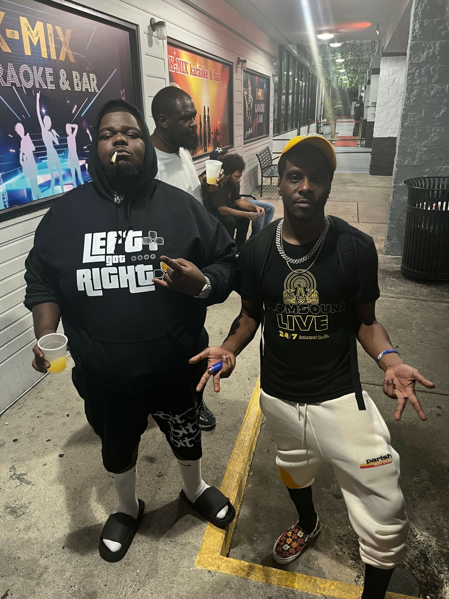 Two people standing in a parking lot at night, one making a peace sign, the other holding a phone, with a store in the background.