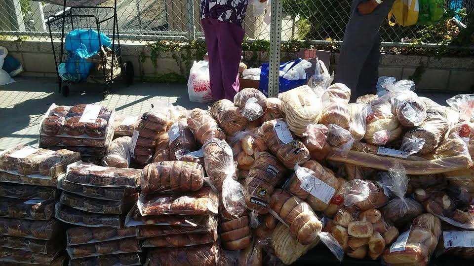 Stacked packages and bags of food on a table outdoors with people in the background.