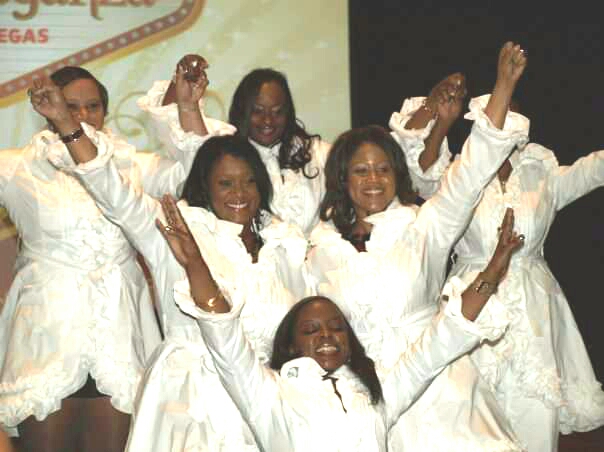 Group of women in white dresses celebrating with raised arms and smiling faces on stage.