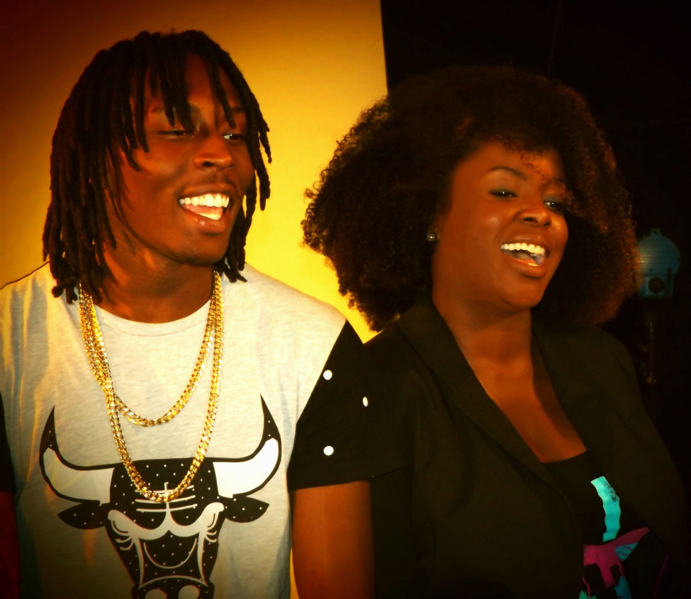 Two people smiling, one with dreadlocks and a white shirt, the other with curly hair and a black shirt, in a dimly lit setting.