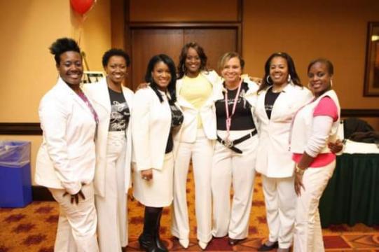 Group of seven women standing together indoors, smiling, dressed in white and pink outfits, with a brown wall background.