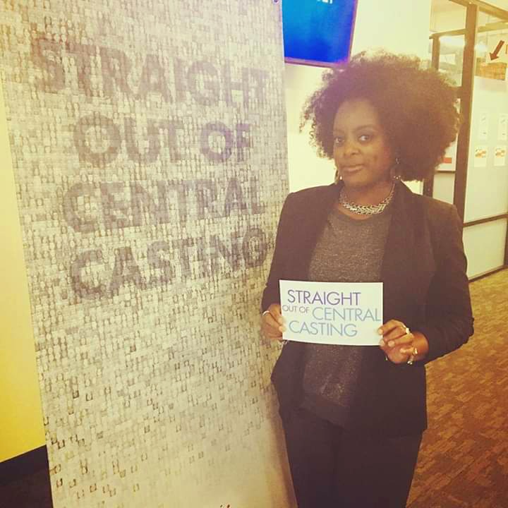 Woman with curly hair holding a sign that reads 'STRAIGHT OUT OF CENTRAL CASTING' next to a textured wall with the same phrase.