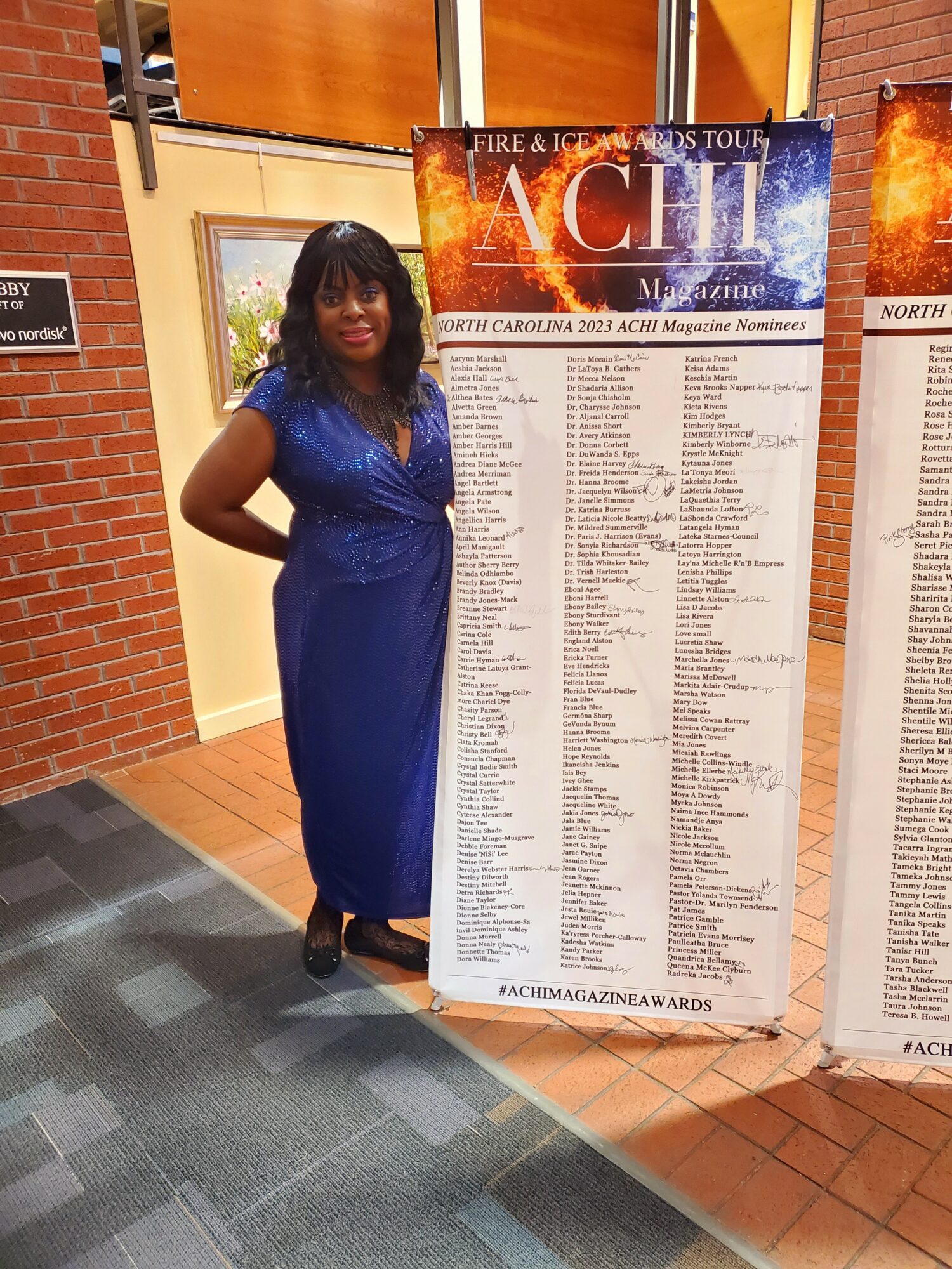 Woman in blue dress standing next to a large event poster, smiling, indoors with brick wall and tiled floor.