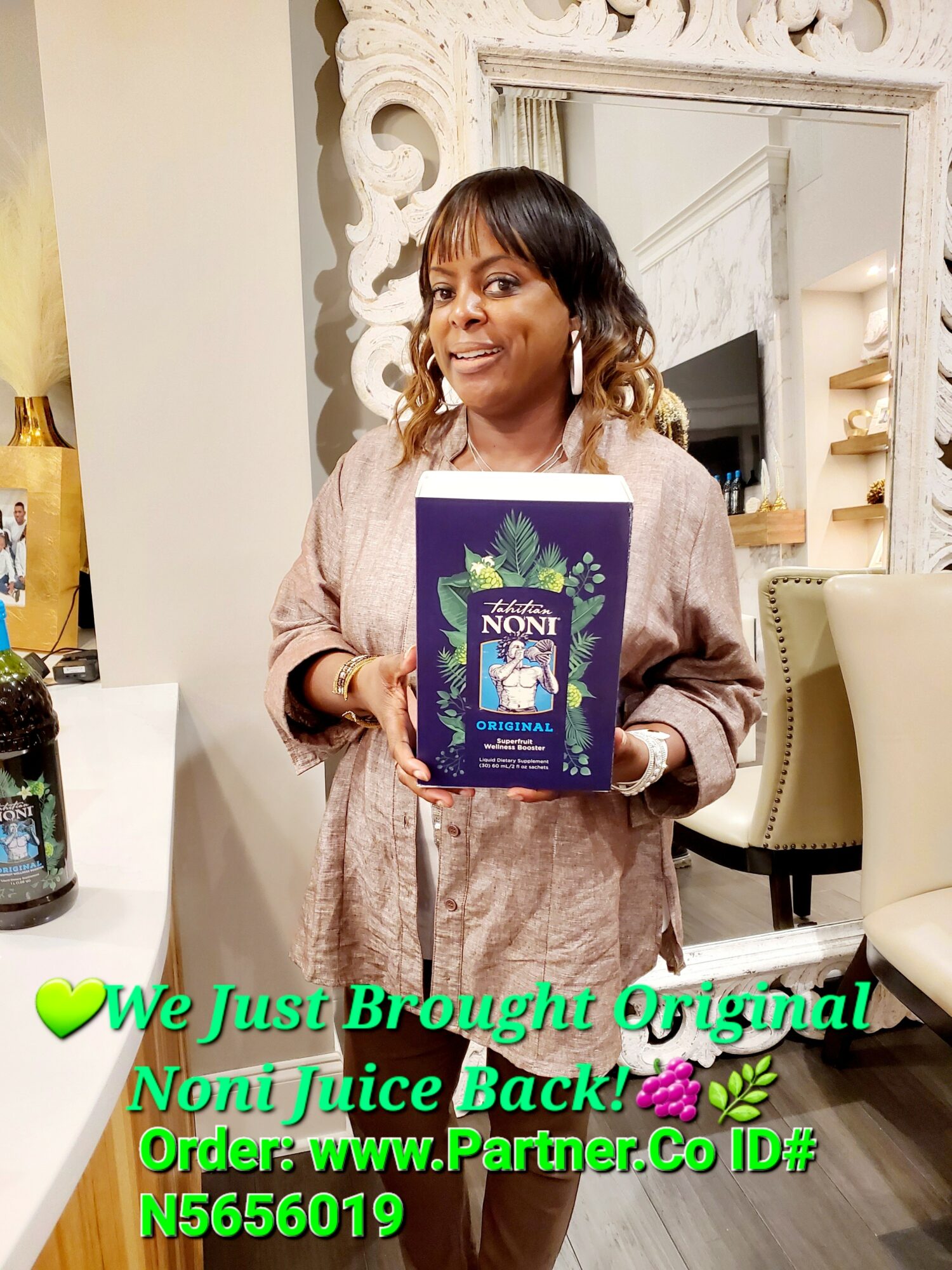 Woman holding a box of Noni juice in a decorated room, smiling, with a mirror and chairs in the background.
