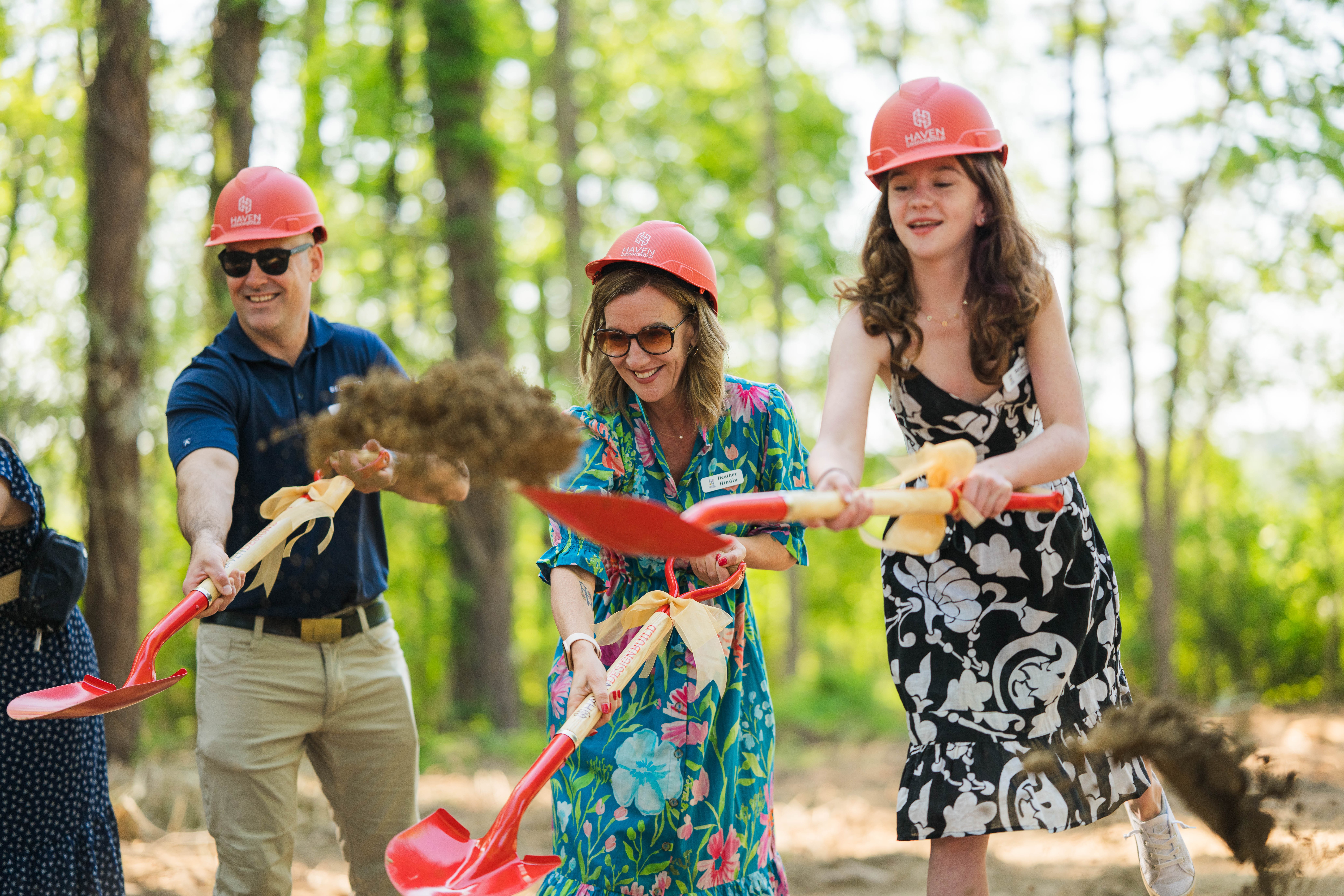 Three children wearing helmets and holding shovels in a forest, smiling and playing together.