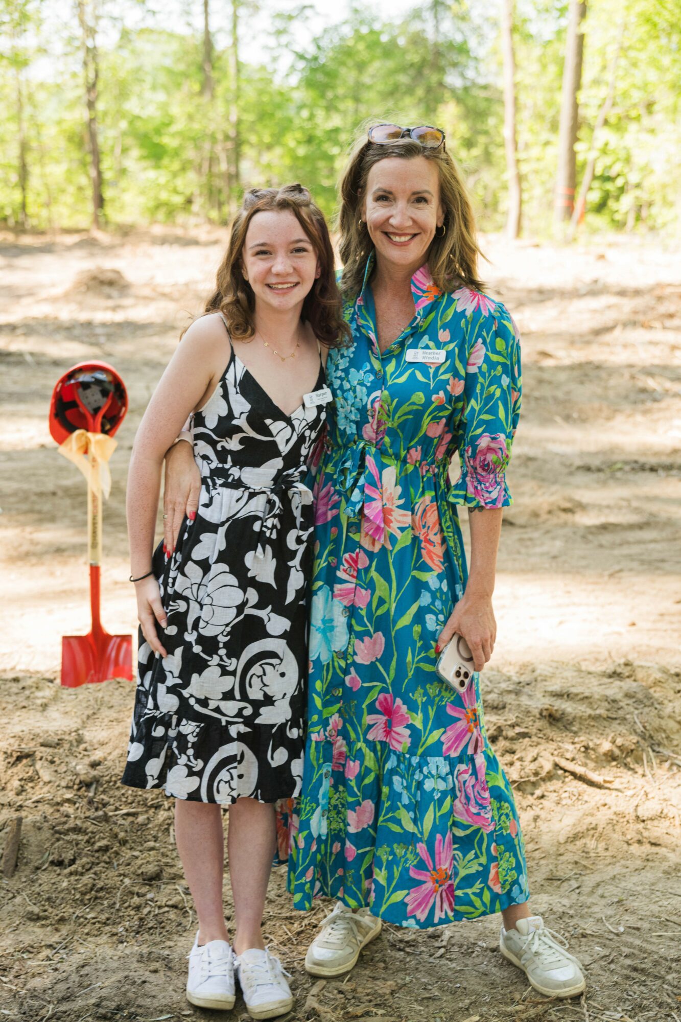 Two women standing outdoors in a wooded area, smiling, one in a black and white dress, the other in a colorful dress.