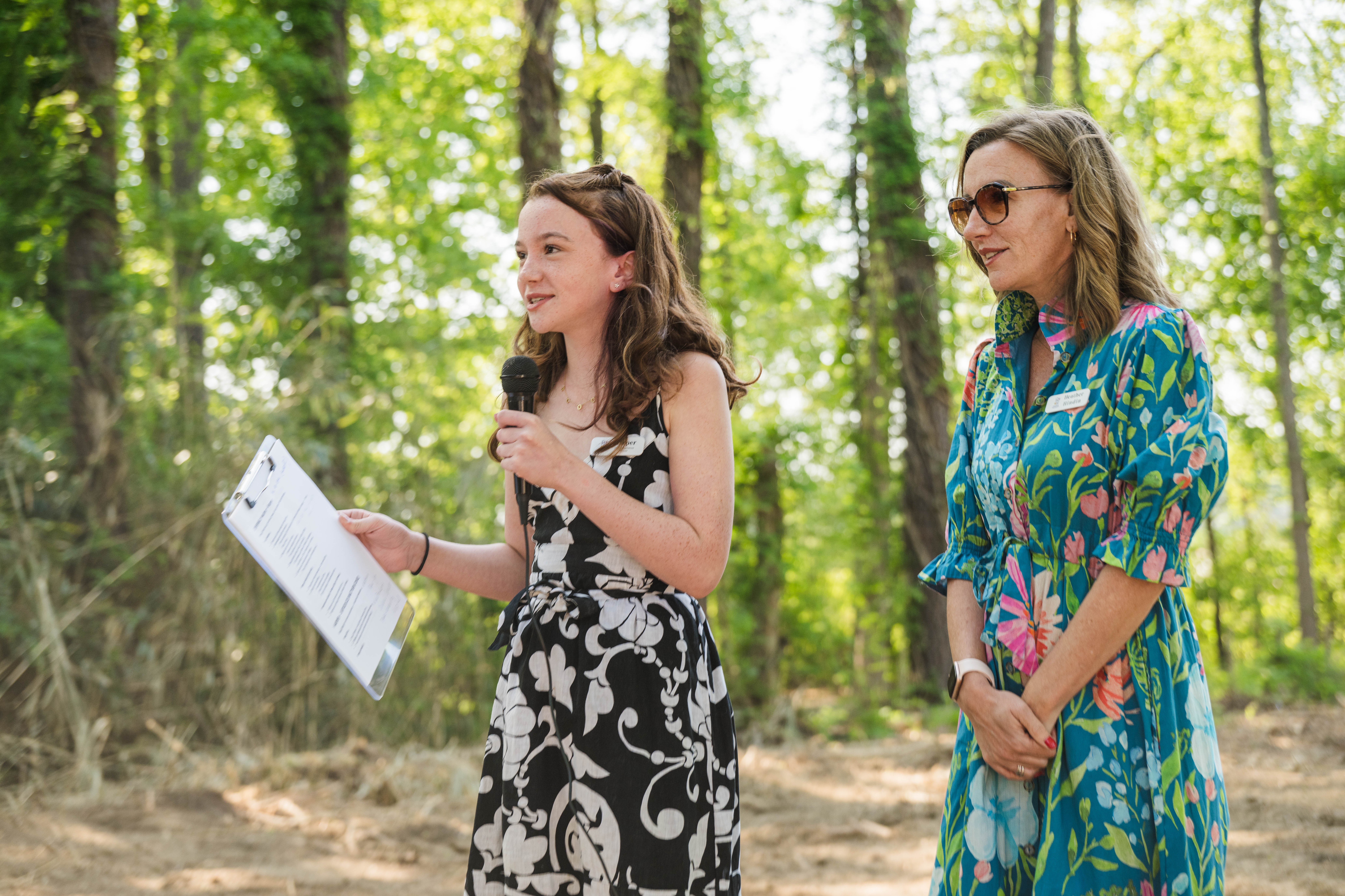 Two women standing outdoors in a forest, one speaking into a microphone and holding papers, the other listening.