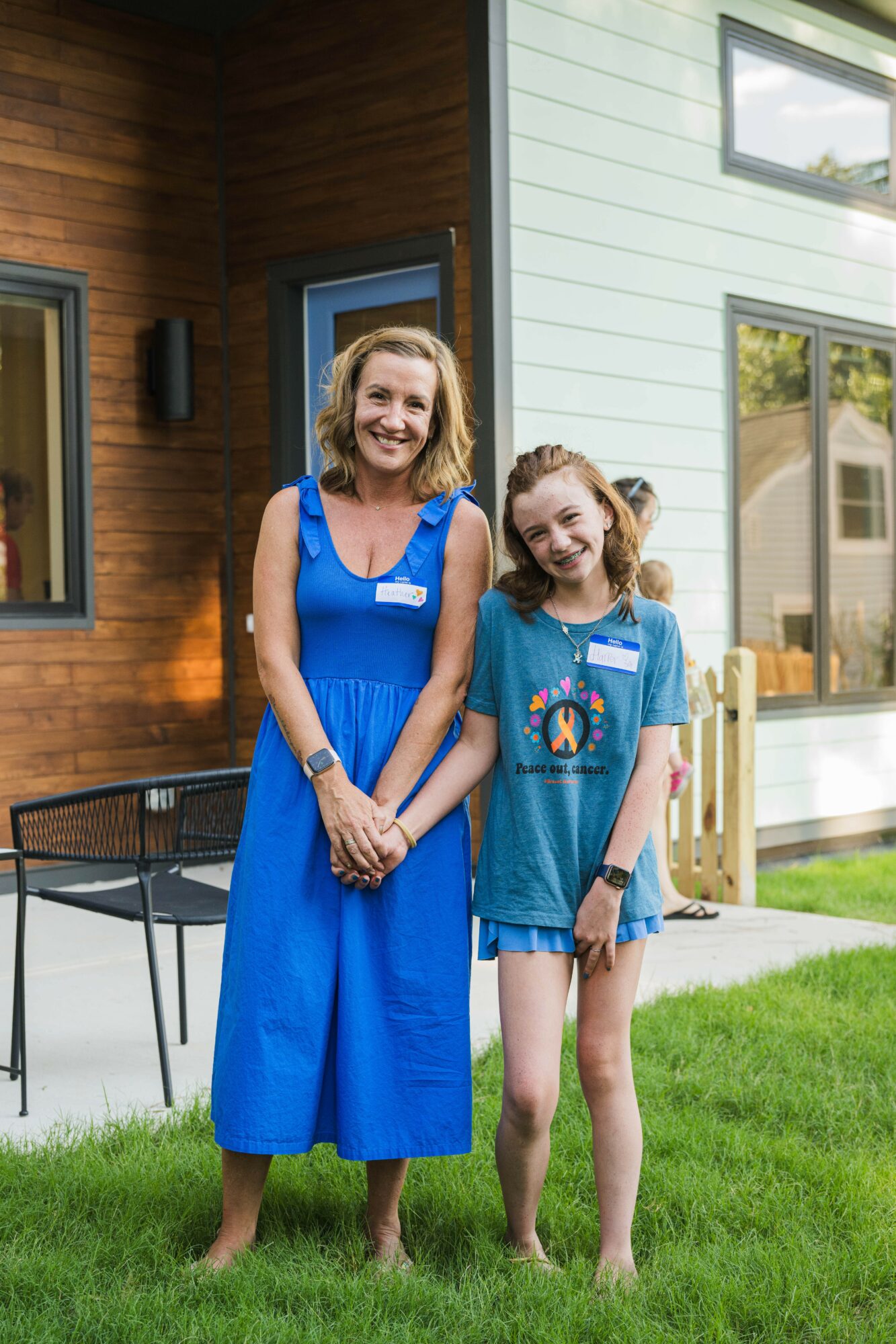 Two smiling girls stand outside a modern house with large windows, grass, and a black outdoor chair nearby.