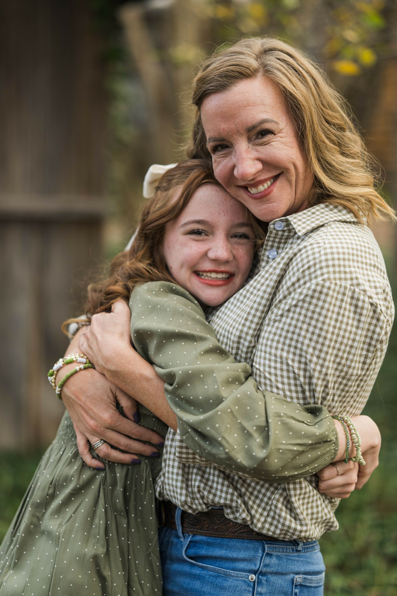Two smiling females hugging outdoors, one appears older with wavy hair, the other younger with straight hair, both wearing casual clothing.