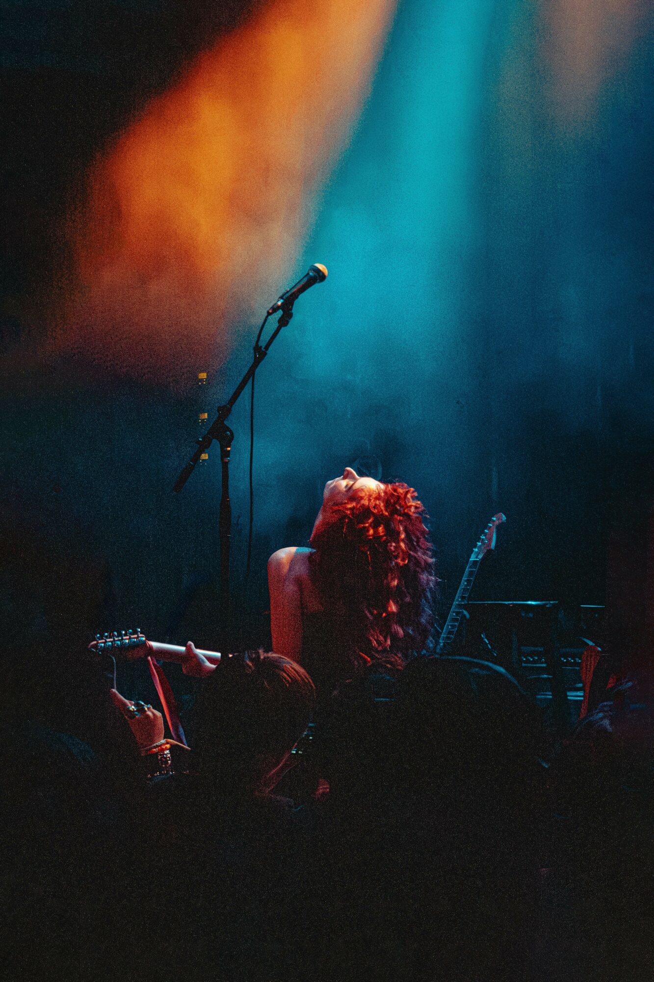 Person with curly hair on stage, holding a guitar, with a microphone stand and colorful stage lighting.