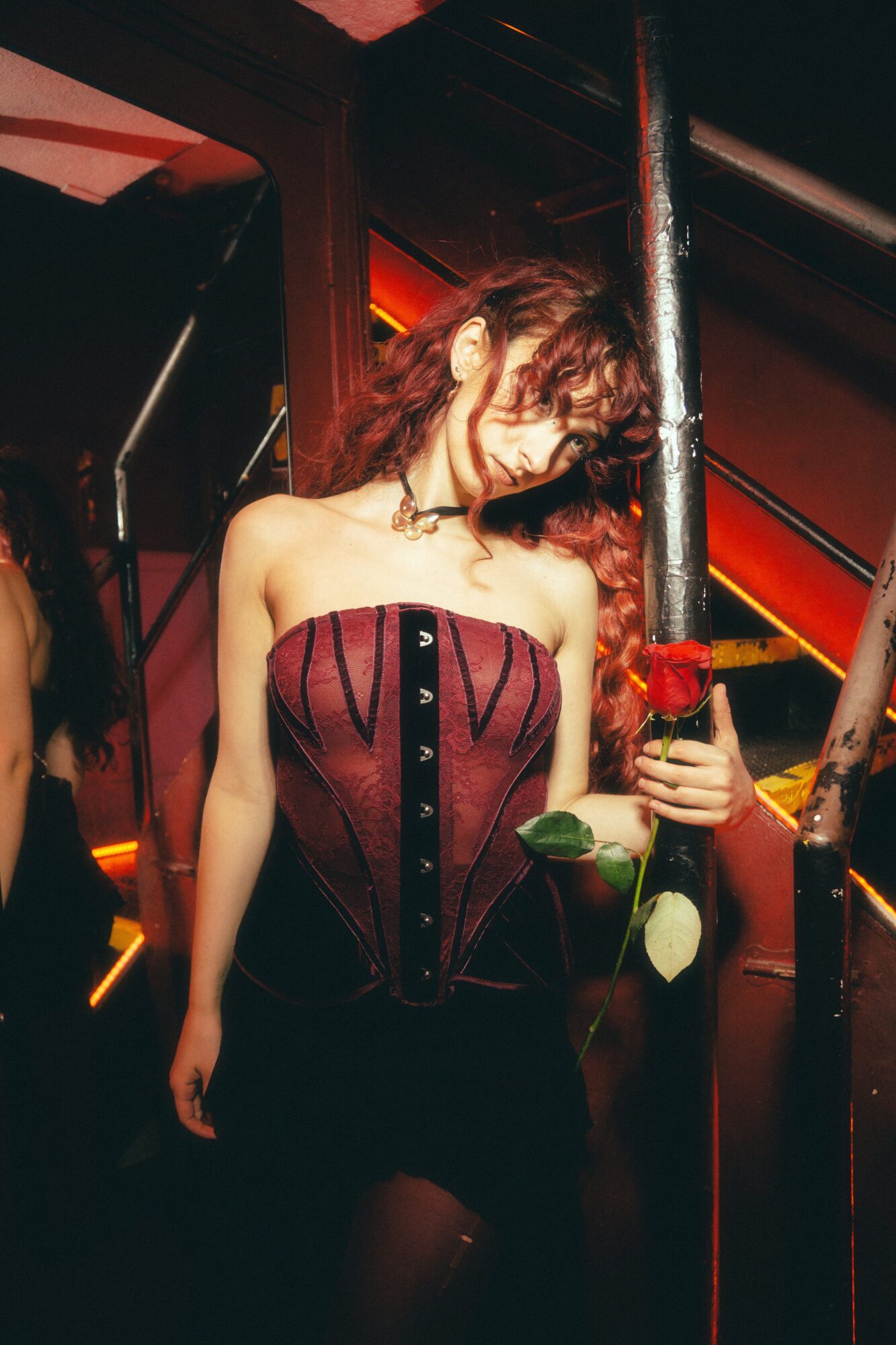 Woman with curly hair in a strapless top holding a flower and a drink, standing in a dimly lit setting.