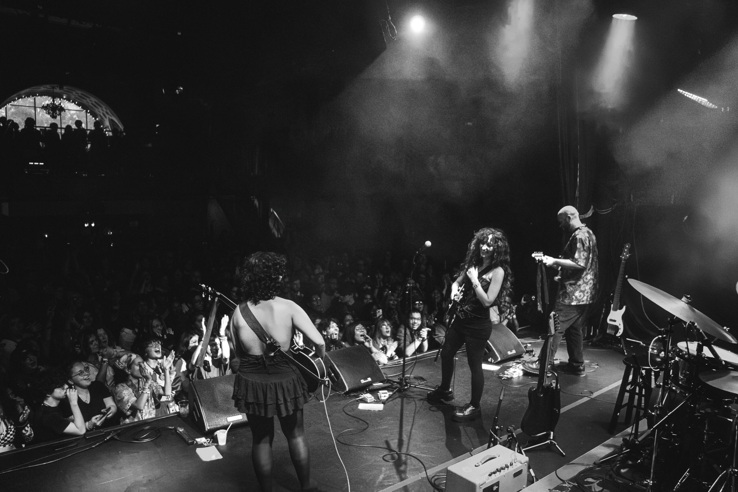 Band performs on stage with audience watching, stage lights shining, in black and white, lively concert scene.