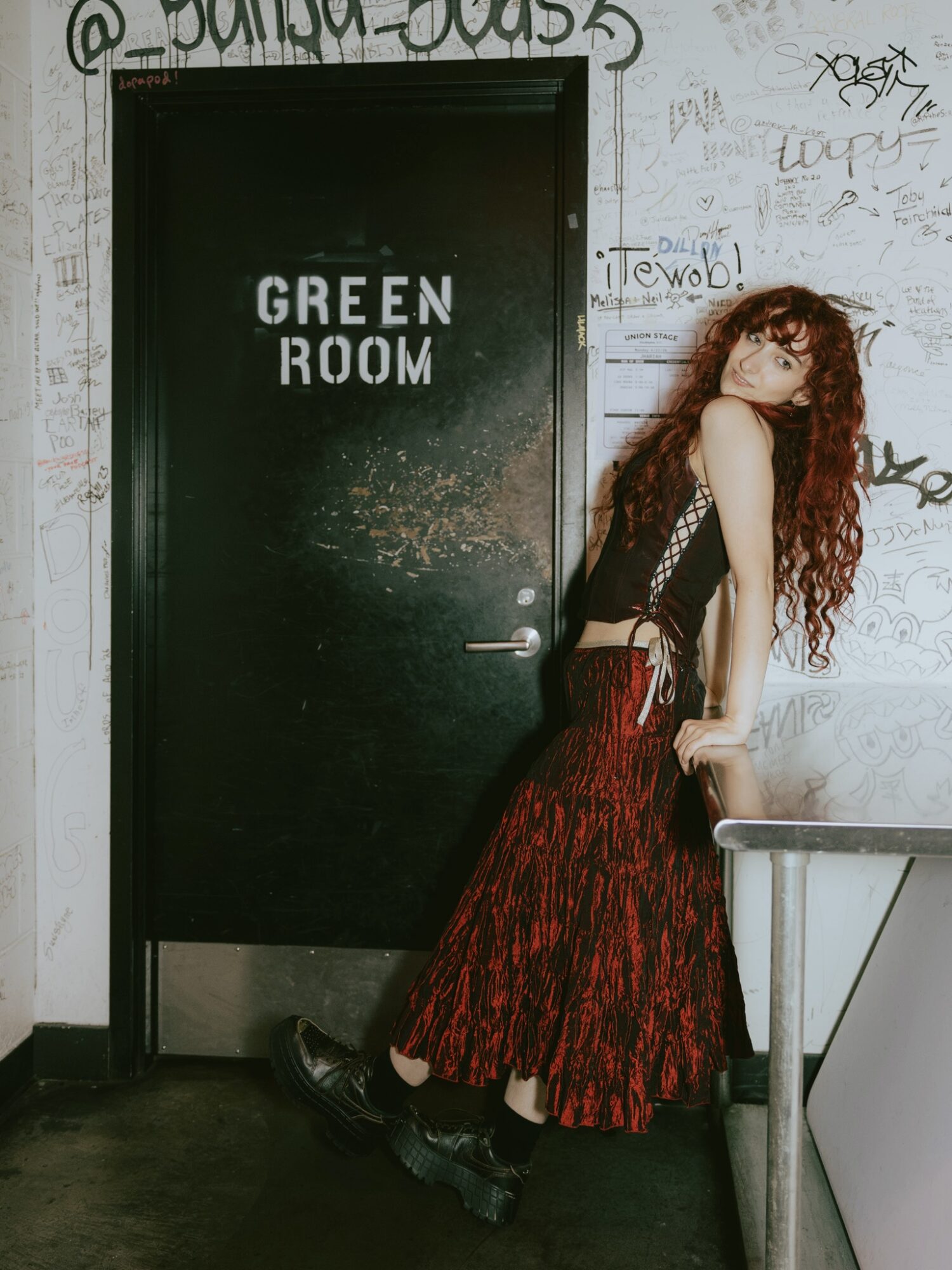 Young woman with long curly hair leaning against a railing in front of a black door labeled 'GREEN ROOM' in a graffiti-covered wall.