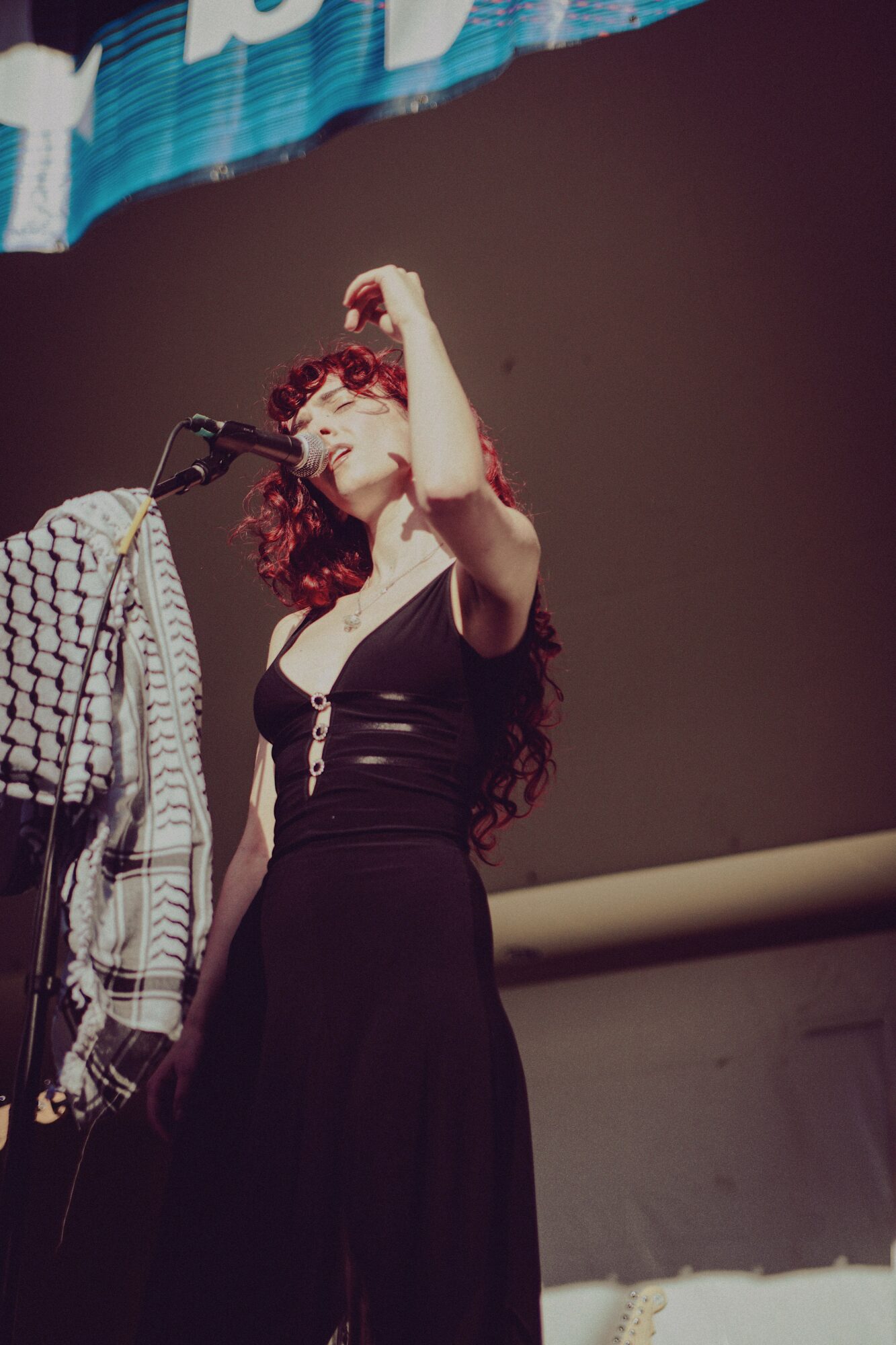 Woman with curly hair and glasses standing on stage, holding her hand up near her head, next to a microphone.