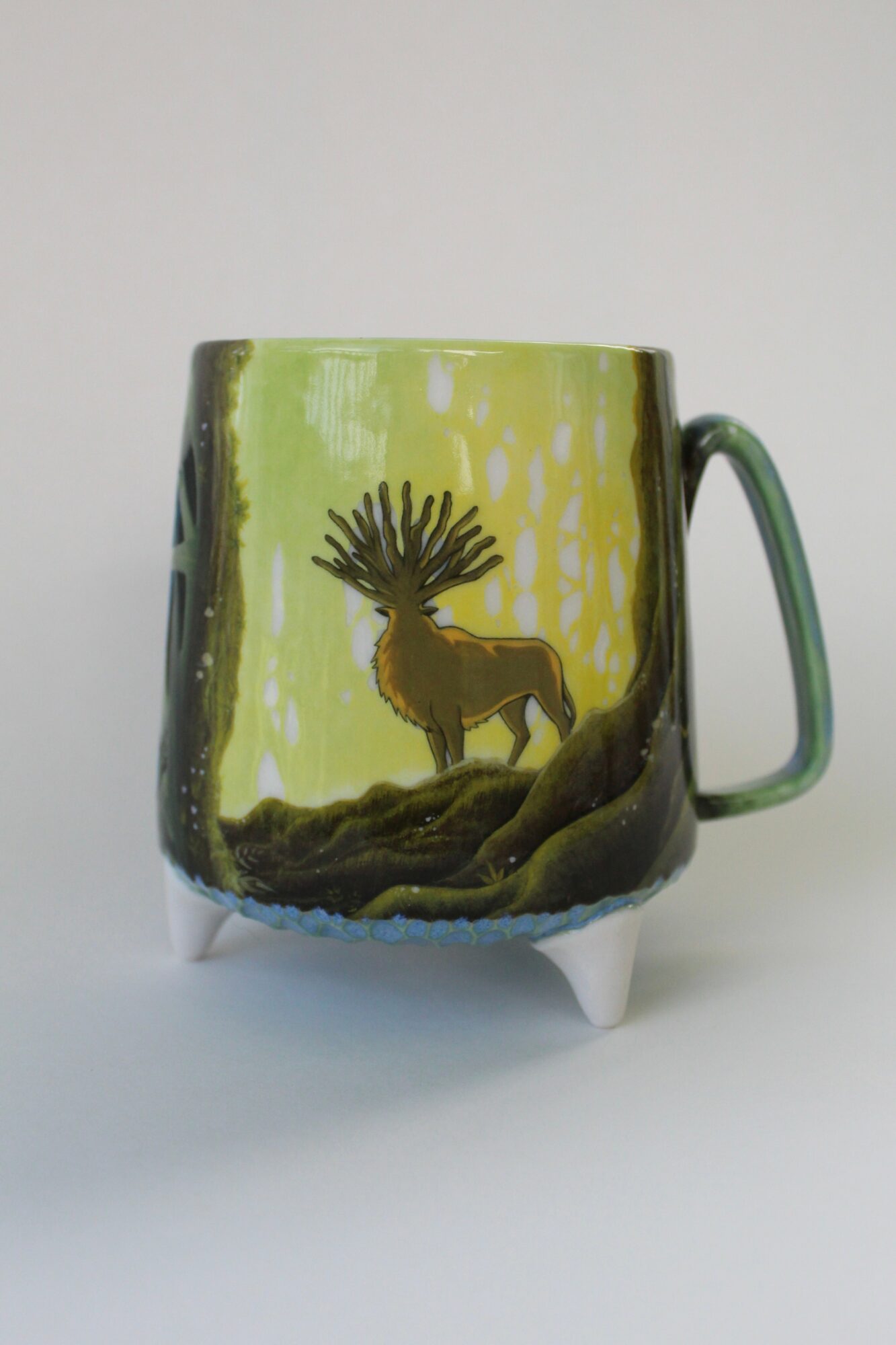 Decorative mug with a deer and forest scene, featuring a tree and ground, on a white background.