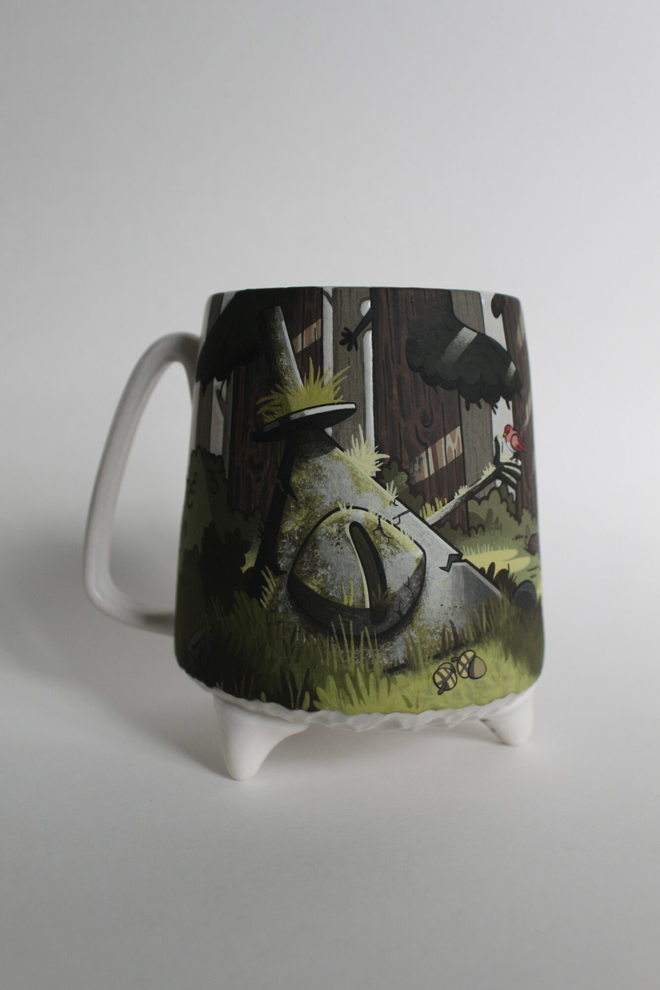 Ceramic mug with a forest scene, trees, rocks, and a small red object, on a plain background.